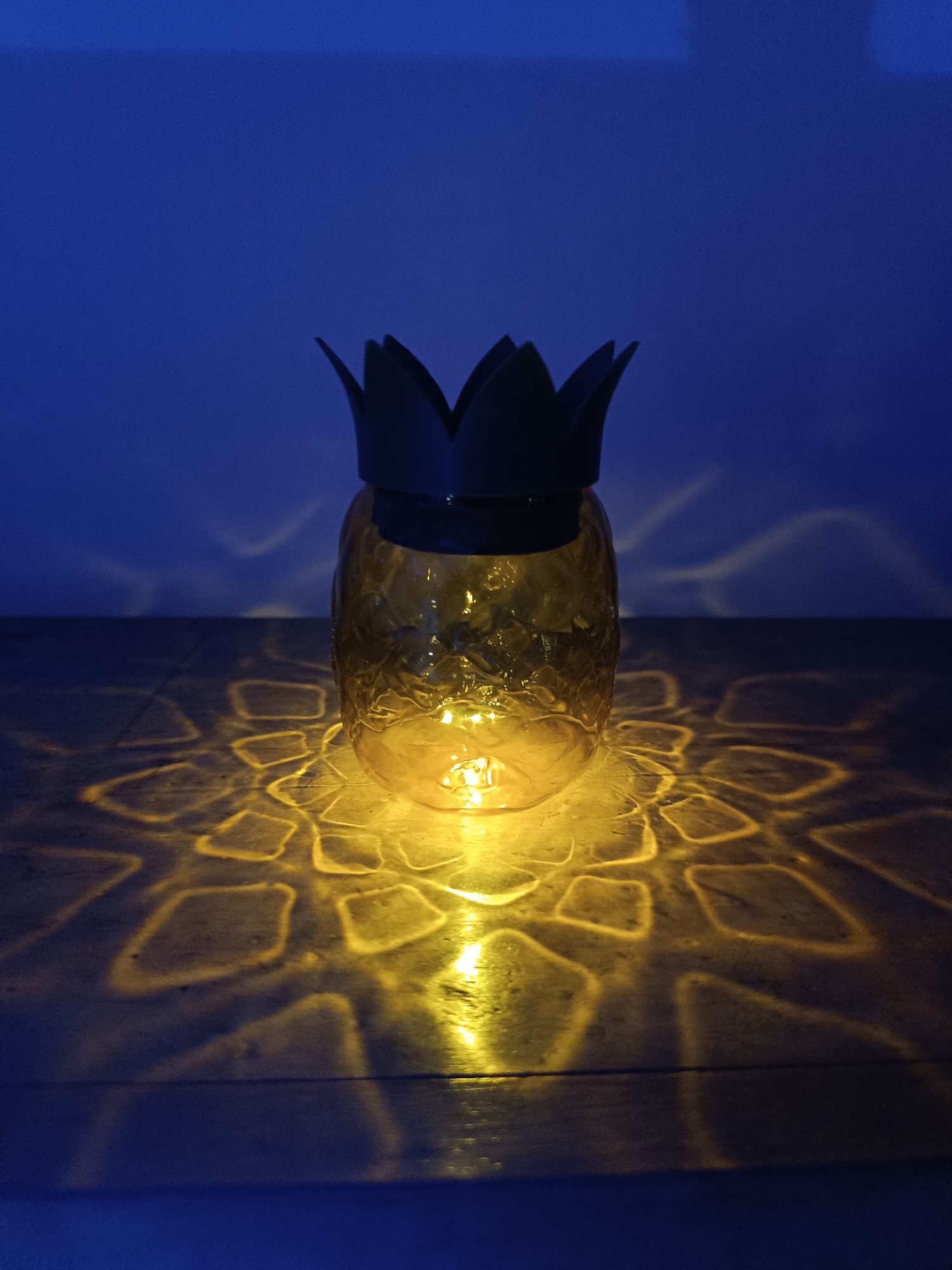 The image depicts a pineapple-shaped lantern placed on a dark surface. The lantern is yellow with a black, leafy crown. A light source inside the lantern projects a patterned, yellow glow onto the surface. The background is a dark blue, likely a wall. The location of the scene within Cherkovitsa, Bulgaria, is not discernible from the image. There are no people present in the image.