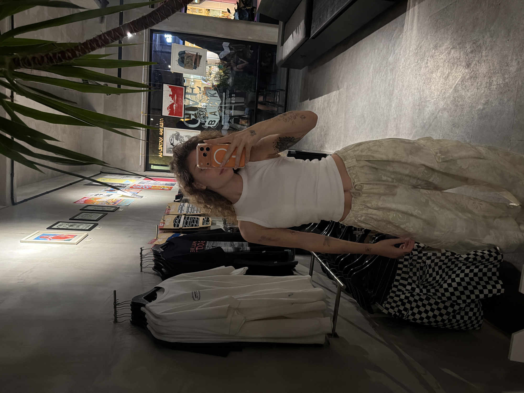 A person is taking a selfie with their phone in a retail store. The person is wearing a white tank top and light-colored patterned pants. They have tattoos on their arm. The store appears to be a clothing boutique or skate shop, with t-shirts, skateboards, and artwork displayed on the walls and racks. The floor is a polished concrete, and there are palm fronds from a large plant in the foreground. The lighting suggests it is nighttime or indoors. Visible text includes words on the t-shirts and artwork, but is not clearly legible. The location context is North Kuta, Indonesia.