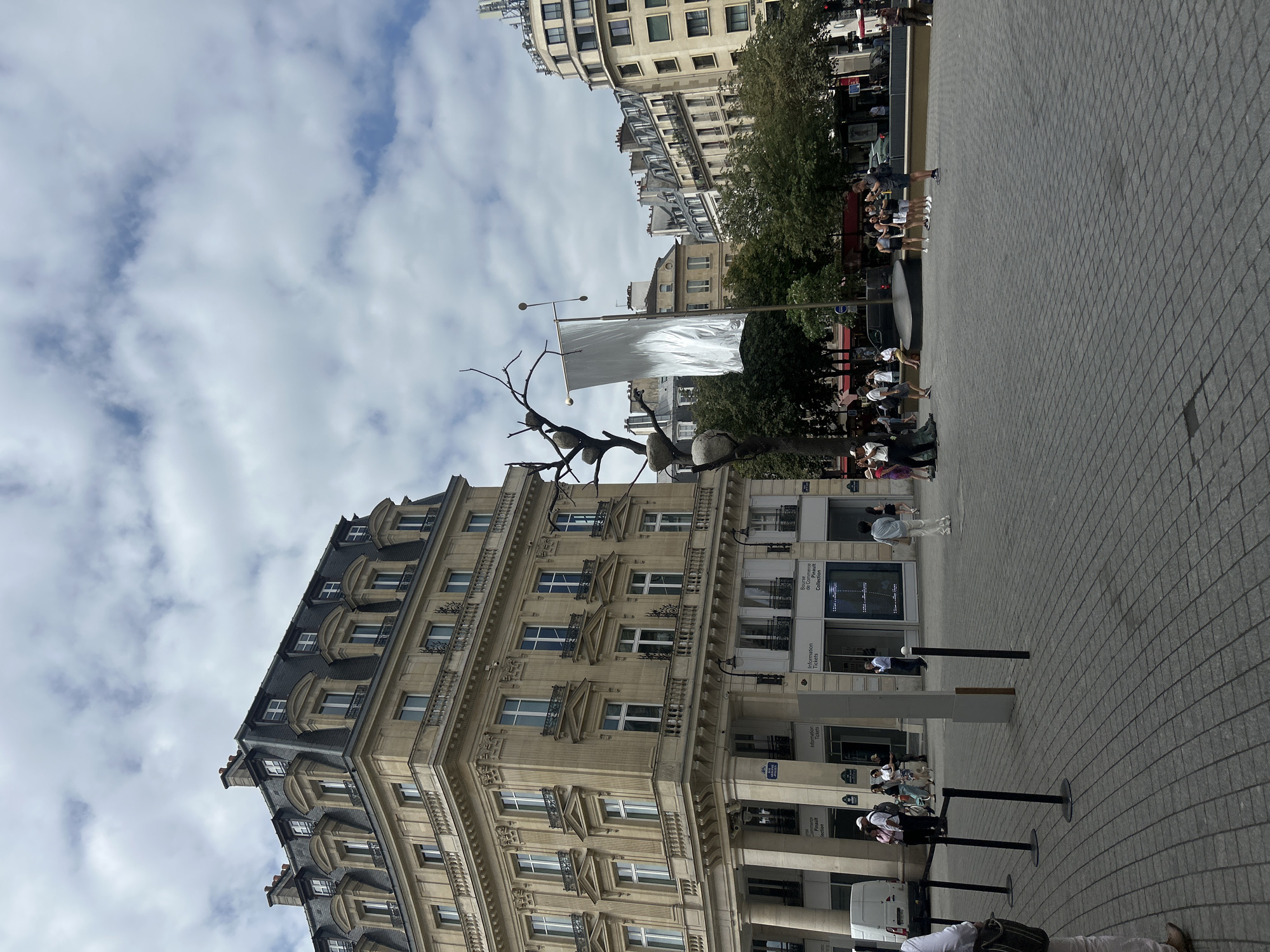 This is a medium shot, eye-level, outdoor photograph of the Place de la Bourse in Paris, France. The image is captured on an overcast day. In the foreground, a paved plaza with a grey cobblestone surface occupies the majority of the frame. Several black stanchions with black ropes are visible on the left side of the plaza, suggesting a designated pathway or waiting area.
To the left, a multi-story building with a light beige facade and a mansard roof with dormer windows dominates the scene. The building features ornate architectural details, including balconies and decorative molding. Below, the ground floor has storefronts with large windows. One storefront displays signs reading "Information Tickets" and "Bourse de Commerce - Pinault Collection." A white van is parked to the far left.
In the center of the plaza stands a contemporary art installation: a stylized tree sculpture made of dark metal, with several large, grey, egg-shaped objects attached to its branches. A large, flowing, silver flag or banner is suspended from a pole next to the sculpture. Several people are scattered throughout the plaza, some walking, others standing and observing the sculpture or the buildings.
In the background, other buildings with similar Parisian architectural styles are visible, including some with mansard roofs and numerous windows. Green trees line the street behind the plaza. The sky is filled with a mix of white and grey clouds.