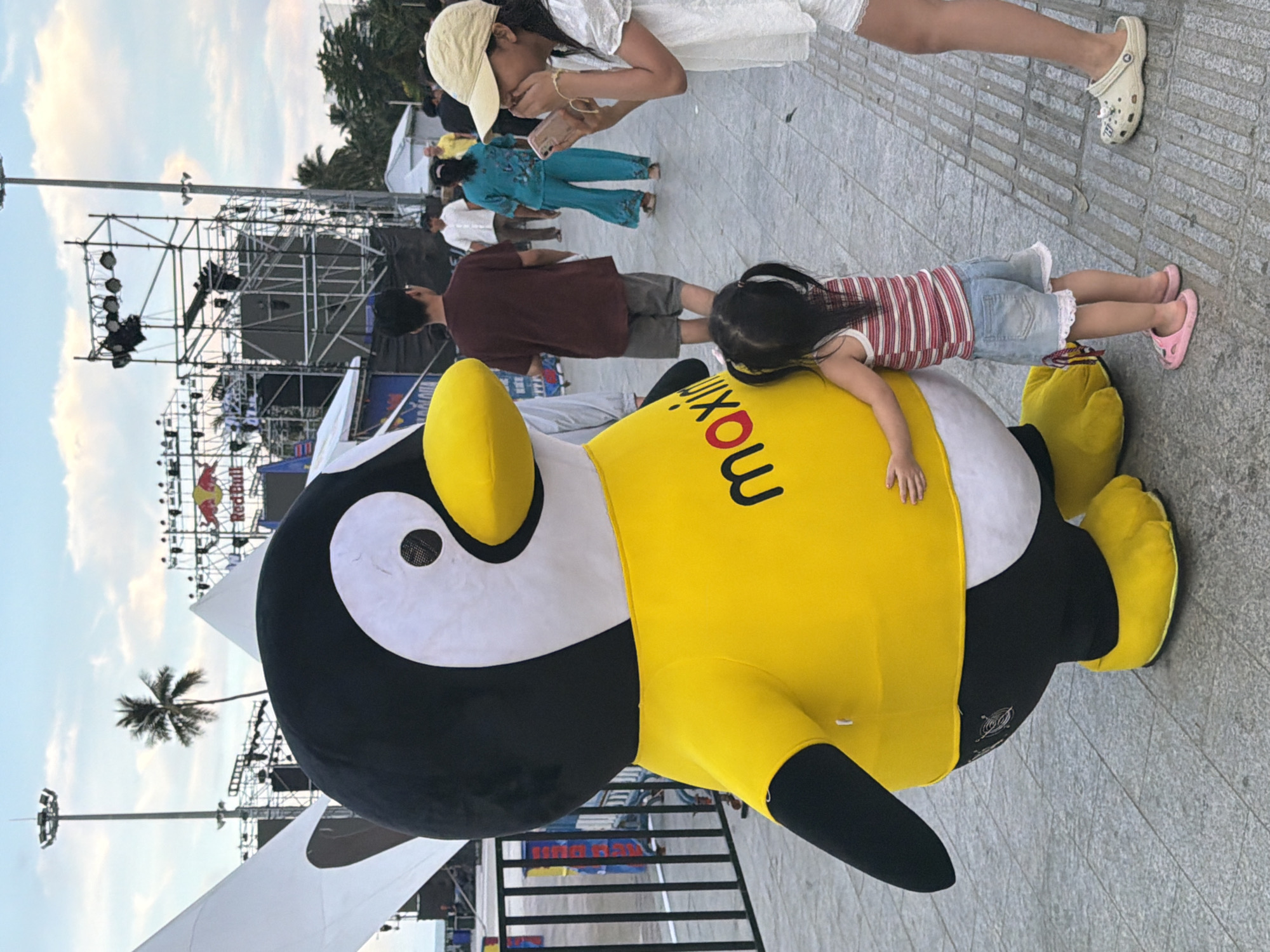 The image depicts an outdoor event scene in Nha Trang Ward, Vietnam. In the foreground, a large inflatable penguin mascot, wearing a yellow shirt with the word "maxim" printed in red, stands prominently. A young girl in a striped tank top and denim shorts hugs the penguin. To the right, a girl wearing a white top and a beige cap is looking at her phone, covering her mouth with her hand. In the background, a crowd of people are visible, along with staging, lighting equipment, and a Red Bull banner, suggesting a festival or performance event. Palm trees and a cloudy sky are visible in the upper portion of the image, indicating an outdoor setting during daylight hours.
