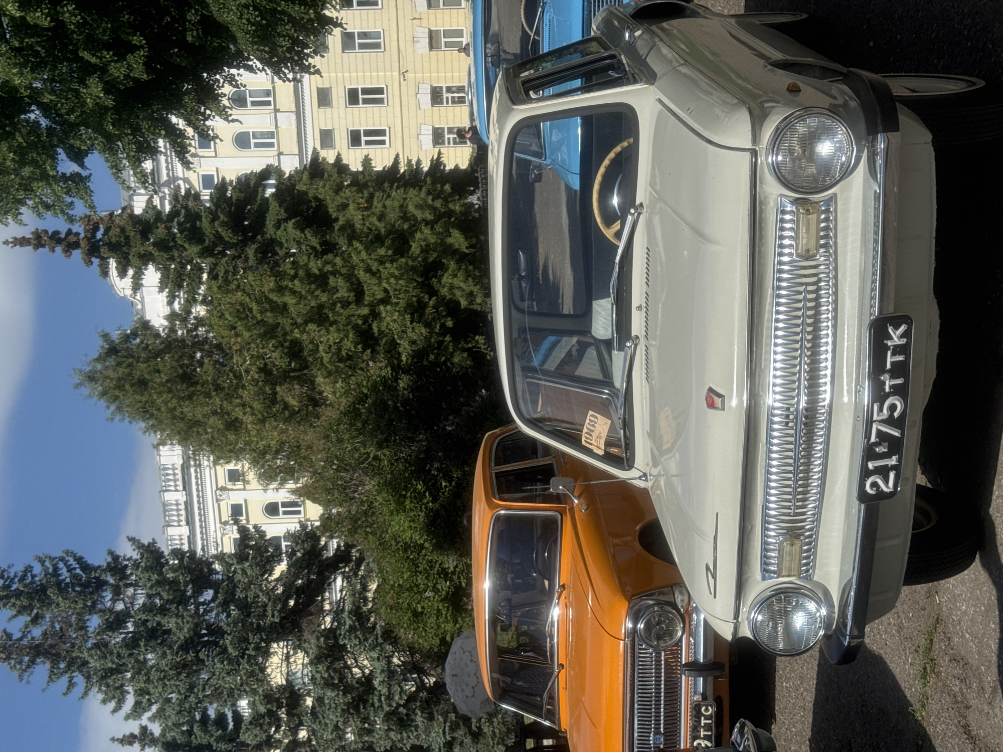 The image shows three vintage cars parked in front of a light yellow building with white trim in Kazan, Russia. The building is obscured by tall, green trees.
The closest car is a light-colored sedan with a chrome grille and a license plate. Next to it, slightly behind, is an orange car with a similar chrome grille. A blue car is partially visible behind the light-colored sedan. The cars are parked on a paved surface. The image is taken on a sunny day with a blue sky.