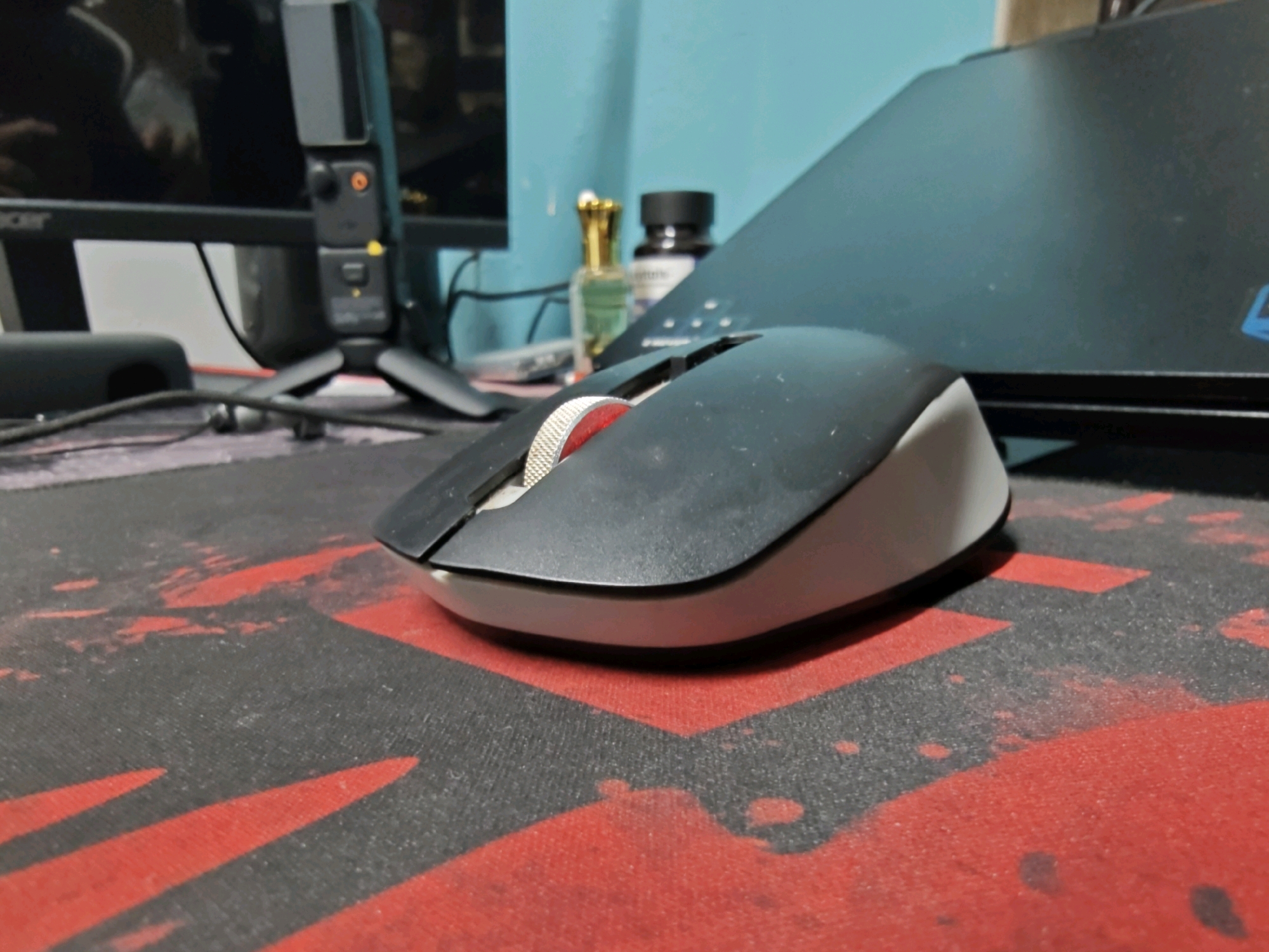 A close-up shot features a wireless computer mouse positioned on a black mousepad with red accents. The mouse is angled towards the right, with its scroll wheel prominently displayed, featuring a metallic band and a red section. The background is blurred, showing a computer monitor on the left and a dark surface, possibly a laptop, on the right. Small bottles of liquid and a dark-capped container are also visible in the background. The lighting suggests an indoor environment. The image does not provide information about the location in the city of Dapoli, India.