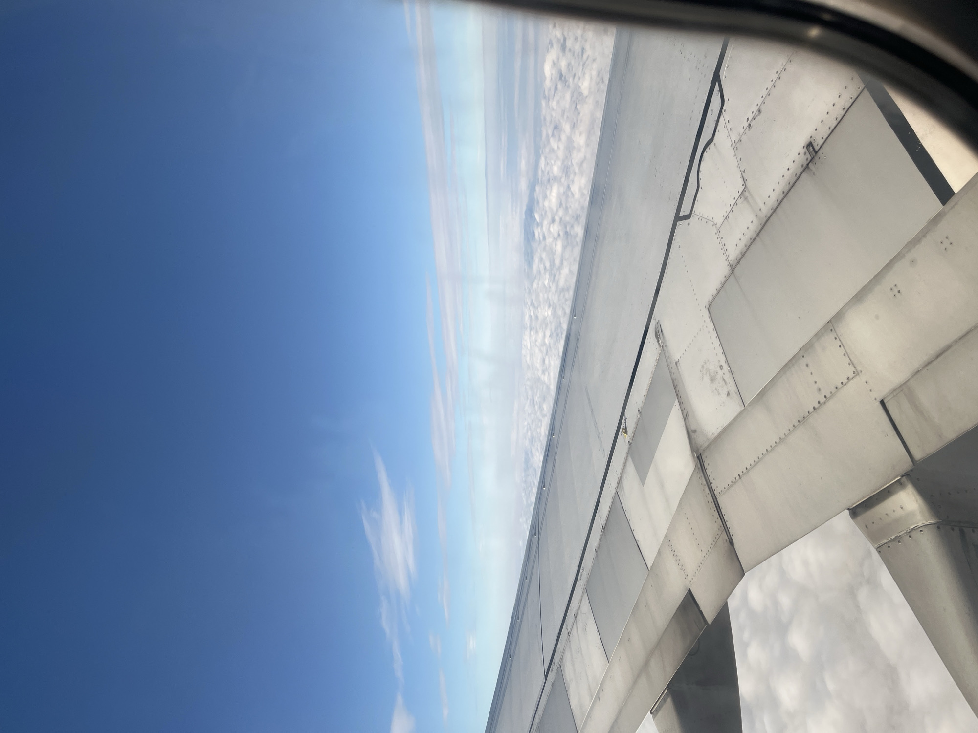 A view from an airplane window shows a vast expanse of blue sky dotted with wispy white clouds. Below the clouds, a thick, white blanket of clouds stretches across the horizon. The metallic surface of an airplane wing occupies the lower half of the frame, with its rivets and panel lines clearly visible. The wing is angled, suggesting the plane is in flight. The scene is devoid of any discernible landmarks or structures that would indicate a specific location within Stolbishche, Russia. There are no people or animals present in the image, and no actions or interactions are depicted.