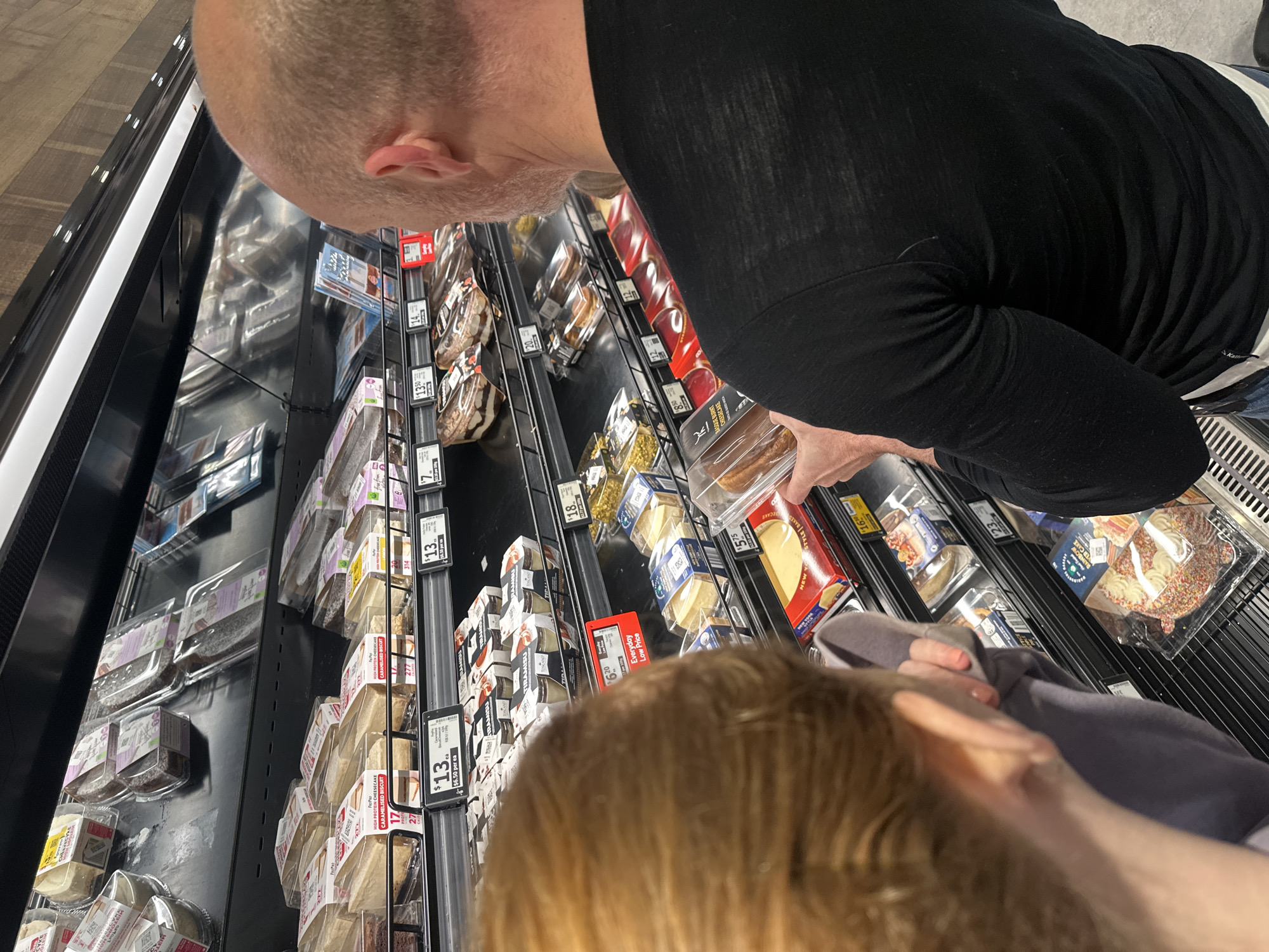 A man and a child are shopping for desserts in a supermarket aisle in Toormina, Australia. The man, bald with a beard, wears a black long-sleeved shirt and is holding a packaged dessert. The child, with reddish-brown hair, wears a grey top and is looking at the desserts. The aisle is stocked with various packaged cakes, cheesecakes, and tiramisu. A shelf below displays cheese products. Price tags are visible on the shelves. The lighting is bright, coming from overhead fluorescent lights.