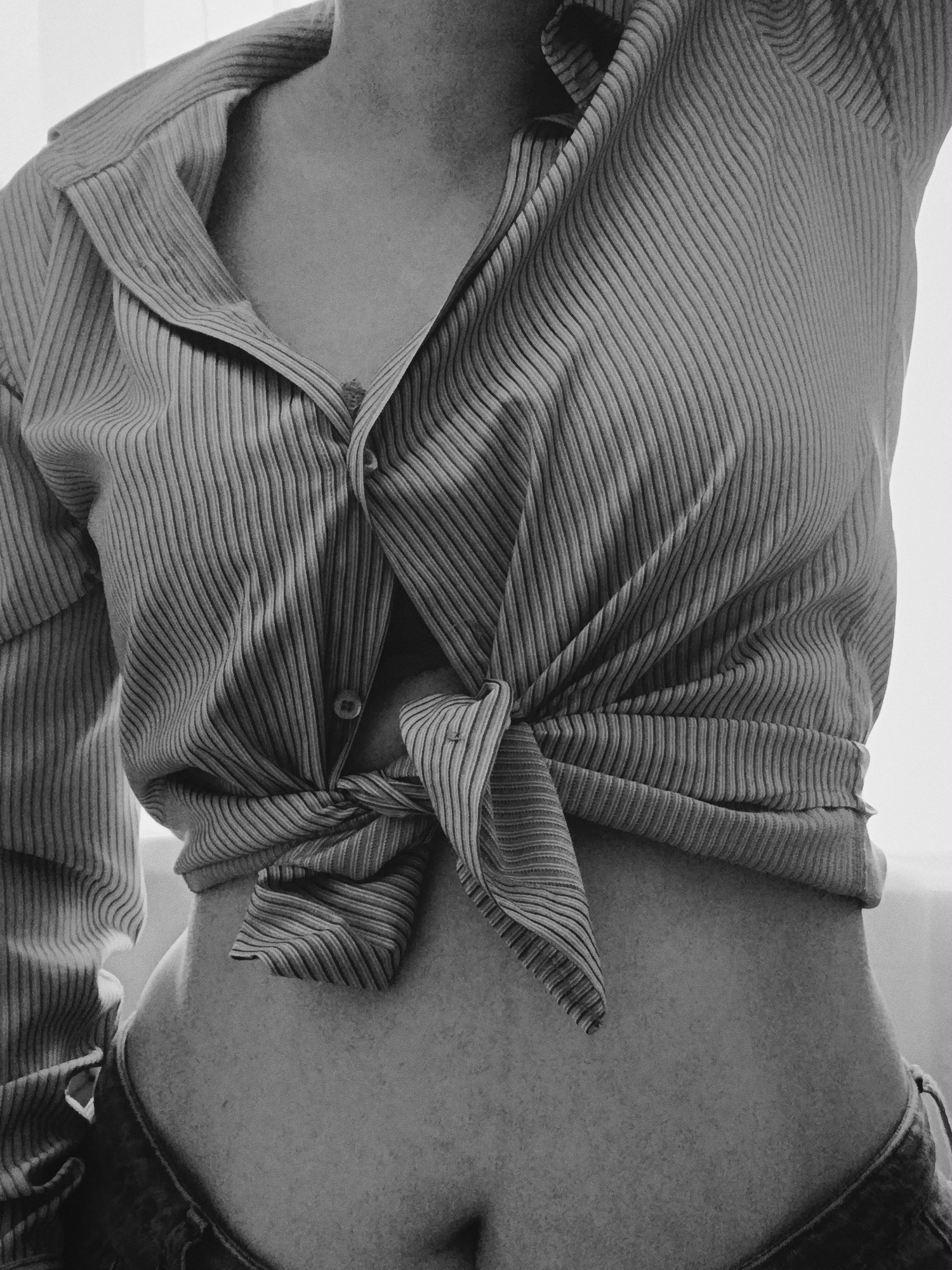 A grayscale, waist-up shot shows a person wearing a striped, long-sleeved shirt tied in a knot at the waist, exposing the abdomen. The shirt is unbuttoned at the top, revealing a small portion of the chest. The person is also wearing jeans, visible just below the waist. The location is not discernible from the image.