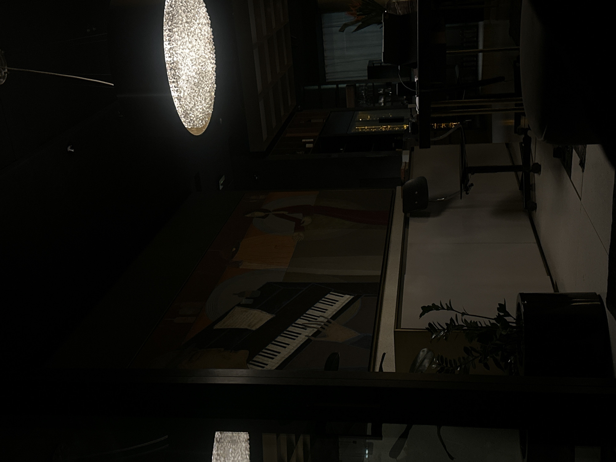 The image depicts a dimly lit interior, likely a restaurant or lounge, with a large, round, illuminated light fixture hanging from the ceiling on the left side of the frame. Below the light, there's a wall with a mural of a piano and what appears to be human figures. To the right of the mural, the floor is visible, showing a gray tiled pattern, and there's a potted plant resting on a surface in the lower right corner. In the background, there's a bar area with shelves of bottles. The image appears to be taken in a modern setting, possibly in Sofia, Bulgaria, but there's no specific indicator in the image confirming the city.