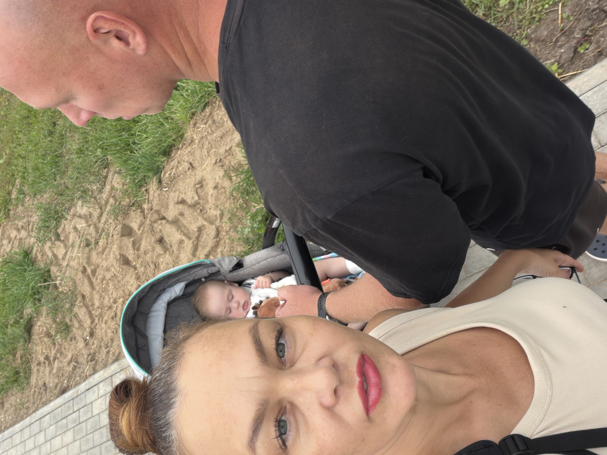 A close-up selfie shows a woman in the foreground with her hair pulled into a bun, wearing a light-colored tank top. To her right, a baby is visible in a stroller. Behind the woman and baby, a bald man wearing a black t-shirt is in profile, facing away from the camera. The background consists of a paved walkway, sandy ground, and green grass. The overall scene appears to be an outdoor setting, possibly a park or pathway in the city of Chaikovsky, Russia, given the context.