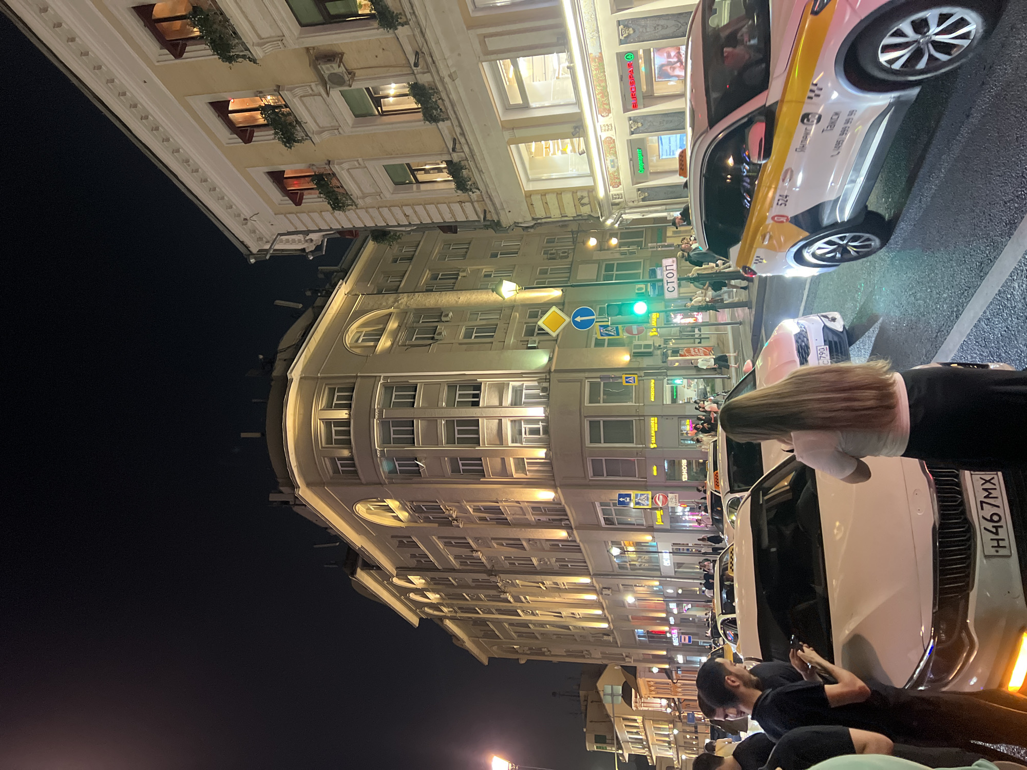 The image shows a nighttime street scene in Moscow, Russia. A prominent, illuminated building with multiple floors and arched windows dominates the left side of the frame. Various shops with lighted storefronts are visible at the ground level of this building, including one labeled "SALAMANDER" and another with Cyrillic text. To the right of this building, another building with yellow facades and red-awninged windows is visible.
Several vehicles are present on the street, including a white car with the license plate H467MX in the foreground, and a white and yellow taxi with the number 524 and Russian text indicating it is a "Yandex.Taxi" service. The taxi has visible signage and contact information. Other cars and taxis are present further down the street.
Several people are standing on the sidewalk and near the cars. One person in the foreground, a woman with long brown hair, is facing away from the camera, looking towards the white car. A man in a black t-shirt is standing next to the white car, looking at his phone. Other pedestrians are visible in the background, walking along the sidewalk.
Traffic signals, including a green light and a stop sign, are visible on poles along the street. The street is paved, and streetlights illuminate the scene. The overall impression is that of a busy urban street at night.
