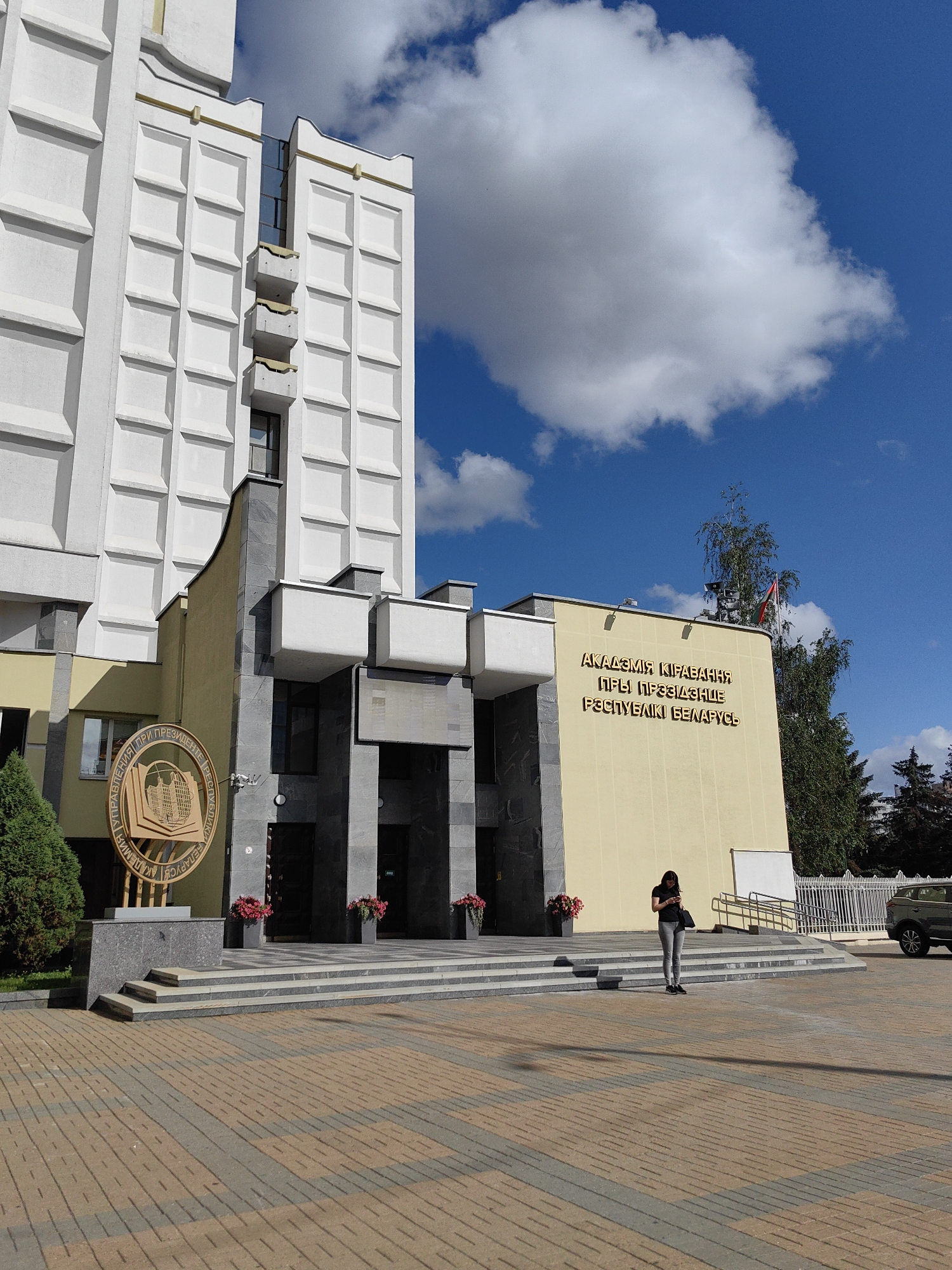 The image captures the exterior of the Academy of Public Administration under the President of the Republic of Belarus, located in Minsk. A woman stands on the paved plaza in front of the building, looking at her phone. The building features a white facade with a distinctive grid pattern, a yellow section with Cyrillic lettering, and a large circular emblem. Steps lead up to the entrance, flanked by planters with red flowers. Trees and a white fence are visible in the background, along with a white SUV parked to the right. The sky is blue with scattered clouds.