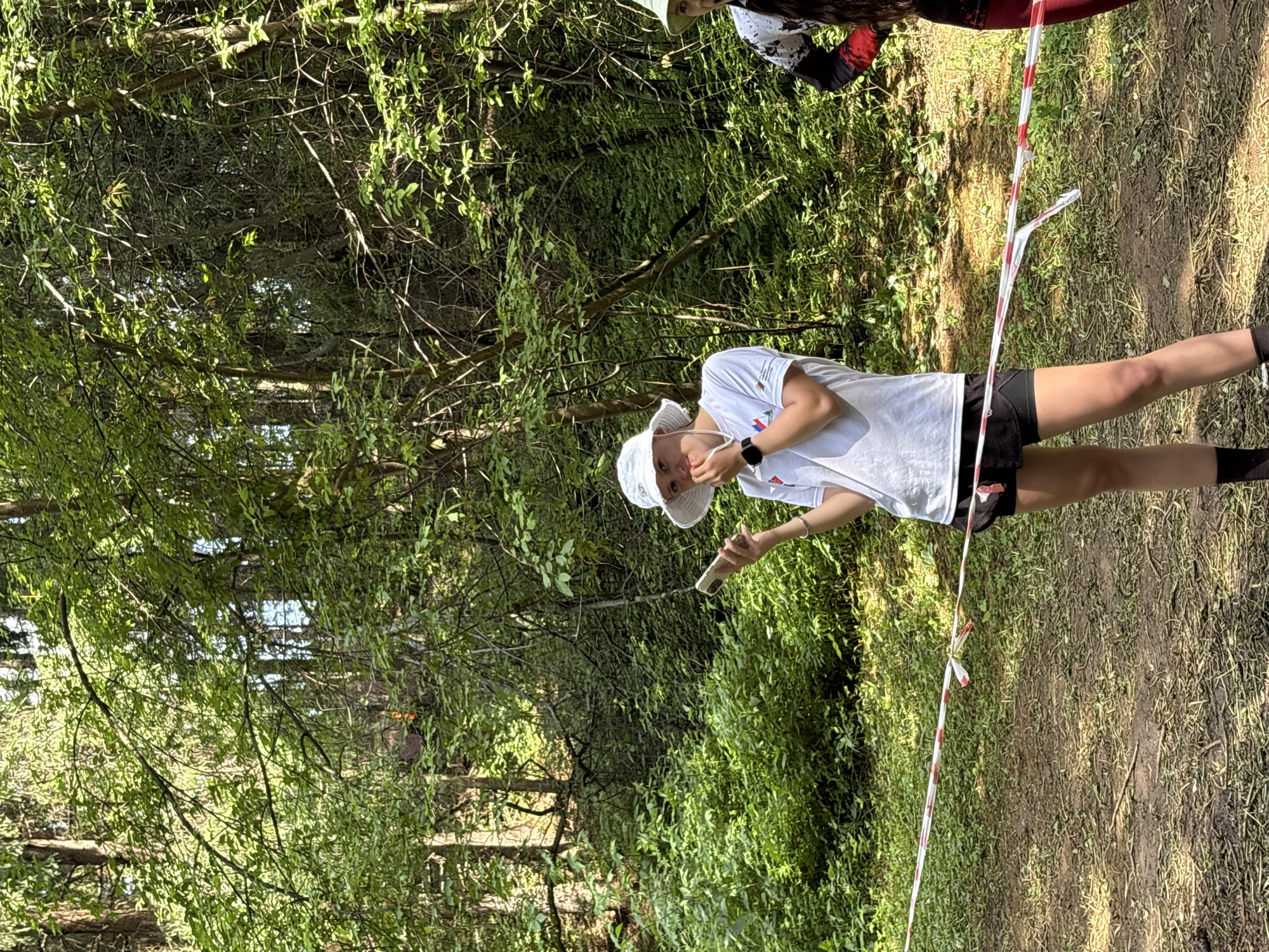 A person wearing a white bucket hat and a white t-shirt stands in a wooded area, holding a white cylindrical object in their right hand. They are wearing black shorts and black socks. A red and white striped ribbon or tape is visible in the foreground, stretching diagonally across the frame. The background is filled with lush green foliage and trees, suggesting a natural outdoor environment. Sunlight filters through the leaves, casting shadows on the ground. Another person, partially visible at the top of the frame, is wearing a dark shirt with red accents and a white bucket hat. The scene appears to be set during the daytime, possibly in a forest or park in Sergino, Russia. The mood seems relaxed and casual. No visible text is present on the person's clothing or the objects.