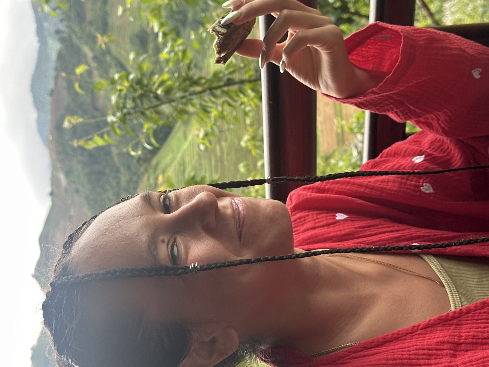 A young adult female is depicted outdoors, smiling and looking towards her right, in what is identified as Púng Luông, Vietnam. She has dark hair styled in numerous thin braids, some adorned with small silver-toned beads. A mole is visible above her right eyebrow and another near her right eye. She is wearing a textured, loose-fitting red garment with small white heart embroideries, over a ribbed, light olive green top. A delicate gold chain necklace is visible. In her right hand, she holds a partially consumed, dark brown, textured food item, possibly a wafer or biscuit, with a light-colored filling or topping. Her fingernails are long, manicured, and have a light, iridescent finish. She is positioned behind a dark brown or red railing, which extends horizontally across the mid-ground. The background consists of out-of-focus lush green foliage, trees, and rolling mountains, some showing signs of terraced cultivation, under a bright, overcast sky.