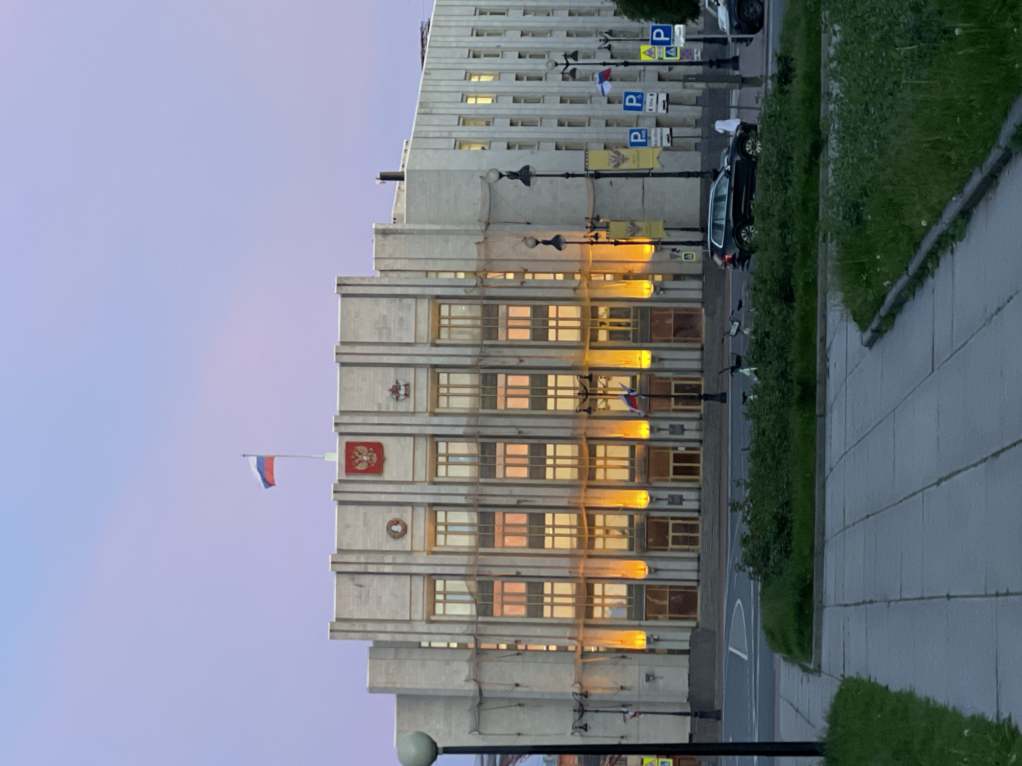 The image shows the State Hermitage Museum, formerly the Winter Palace, located in Saint Petersburg, Russia, during twilight. The building is a grand structure with a light-colored facade, featuring many windows and vertical architectural elements. A Russian flag is prominently displayed on a flagpole atop the building, along with the Russian coat of arms. The building is illuminated by warm lights, casting a glow on its windows and highlighting the architectural details. In front of the building, there is a street with cars parked and driving. A sidewalk and a green space with grass and bushes are visible in the foreground. The sky is a soft gradient of pink and purple, typical of a twilight sky. The overall scene depicts a majestic building in an urban setting as day transitions into night.