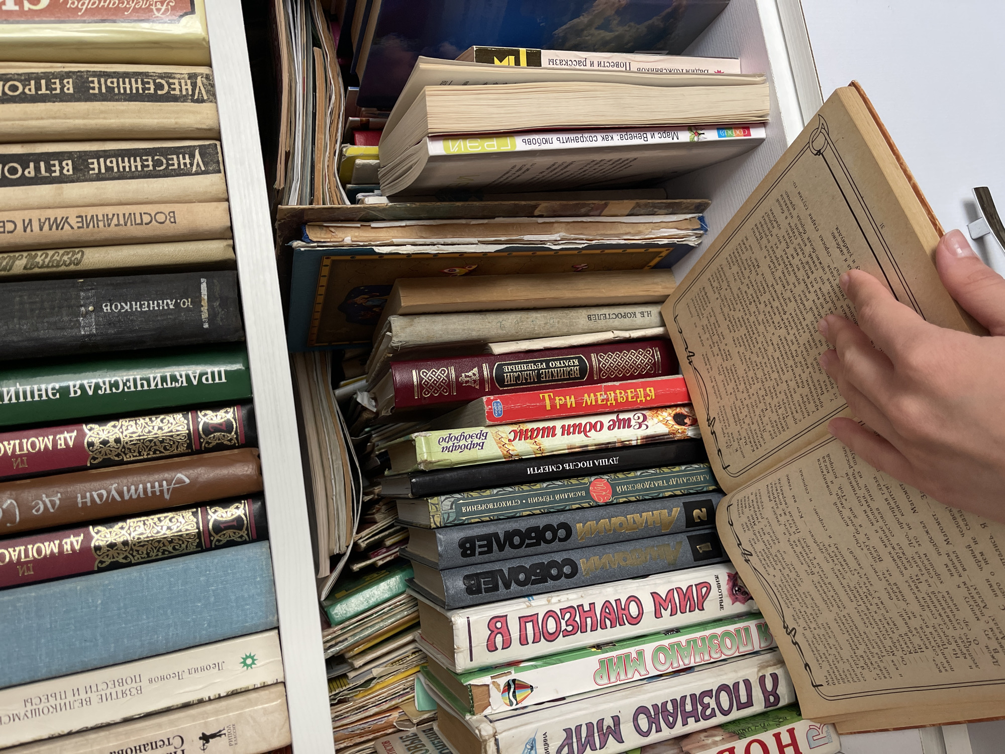 The image shows a close-up, overhead view of a shelf filled with books, predominantly in Russian. A person's hand is visible, holding open a large, old book, its pages filled with dense text and decorative borders. The book is aged, with yellowed pages and a worn cover.
The shelf is packed tightly with books of various sizes, colors, and ages. Some are stacked horizontally, while others are placed vertically. The spines of many books are visible, displaying titles and author names in Cyrillic script. Among the visible titles are "Я ПОЗНАЮ МИР" (I Know the World), "ВЗЯТИЕ ВЕЛИКОШУМСЬ: ПОВЕСТИ И ПЬЕСЫ" (The Taking of Velikoshumsk: Stories and Plays) by Leonid Leonov, "УНЕСЕННЫЕ ВЕТРОМ" (Gone with the Wind), and titles related to history, science, and literature. The books exhibit a range of conditions, from new-looking to quite old and worn.
The overall setting appears to be a personal collection or a bookstore, given the sheer volume and variety of books. The lighting suggests an indoor environment, possibly a dimly lit room or a shelf in a library or home. The focus is on the books and the act of reading or browsing them. The presence of the hand suggests a moment of engagement with the material. The image evokes a sense of discovery and the depth of knowledge contained within the pages.