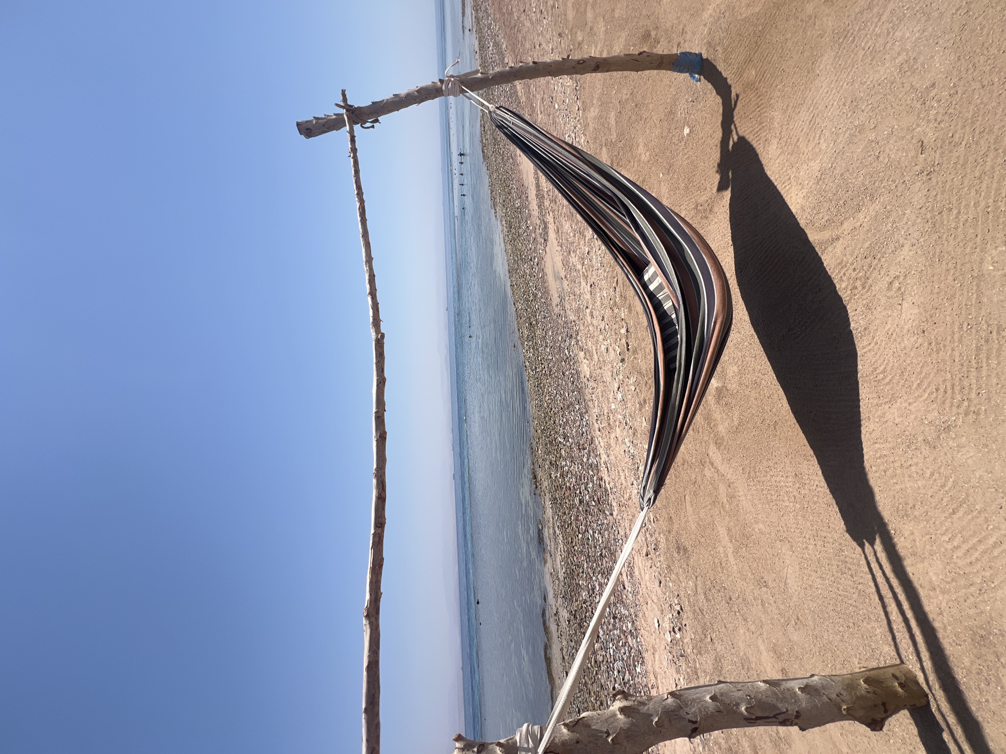 A striped hammock hangs between two wooden posts on a sandy beach in Nuweiba, Egypt. The hammock is made of fabric with horizontal stripes of brown, black, white, and grey. The wooden posts are rough and natural, with ropes securing the hammock to the top of each post. The ground is covered in sand and small pebbles. In the background, the calm, clear blue sea stretches out to the horizon, with a few distant boats and people visible in the water. The sky is a bright, cloudless blue, indicating a sunny day. The image captures a serene beach scene, evoking a sense of relaxation and tranquility.