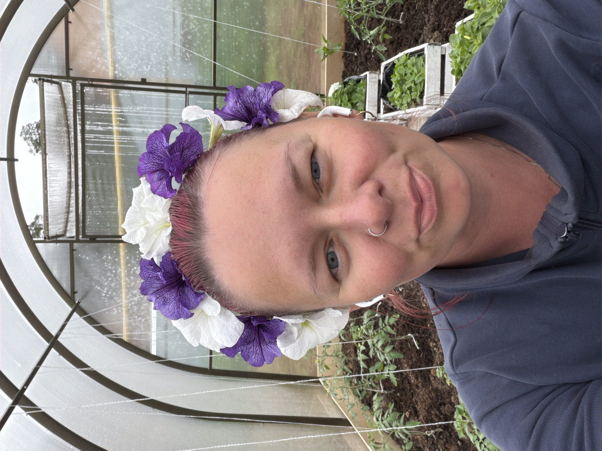 A woman with fair skin and light blue eyes is centered in the frame, wearing a flower crown of alternating purple and white petunias. She has a nose ring and appears to be wearing a gray hooded sweatshirt.
The location is likely a greenhouse, evidenced by the metal frame and translucent covering surrounding the woman. Seedlings in white planters and small plants in the soil are visible in the background, further suggesting a greenhouse environment. The exact location within the city of Chaikovsky, Russia is not determinable from the visual information provided.