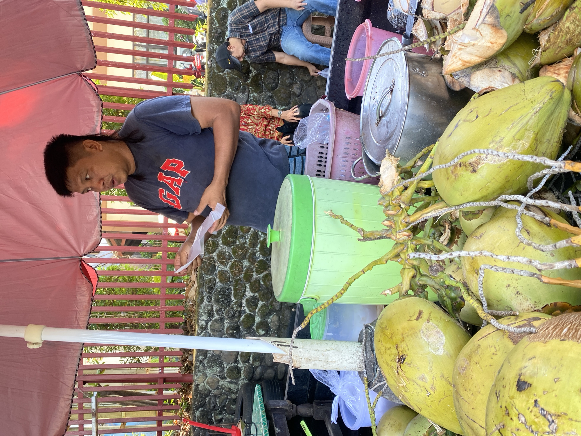 An outdoor scene in Manado, Indonesia, features a man in a dark blue t-shirt with a red "GAP" logo standing under a pink umbrella. He holds a piece of paper in his right hand and appears to be speaking. In front of him, a large pile of green coconuts is visible, some with their husks partially removed. To his right, a green cooler with a lid is positioned next to a clear plastic container. Further to the right, another man wearing a plaid shirt and jeans is seated on a stool, reaching for something in a pink basket. The background shows a stone wall and a red fence with vertical slats.