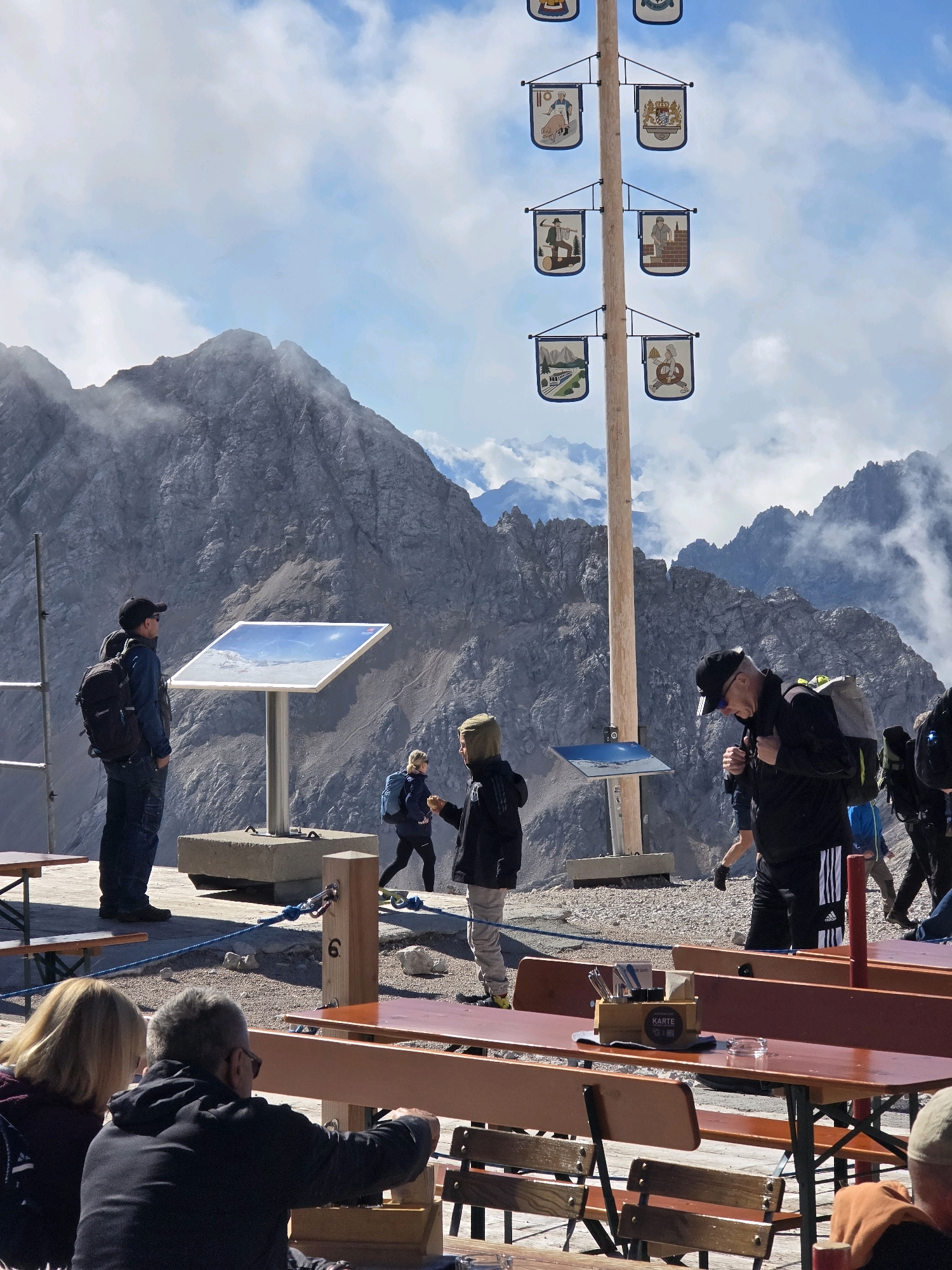 This image captures a scenic view from a high vantage point, likely a mountain summit or viewing platform in the vicinity of Garmisch-Partenkirchen, Germany, given the distinct Alpine landscape. In the foreground, several people are visible, some seated at picnic tables, others standing and observing the surroundings.
One man in a black cap and jacket, wearing a backpack, is looking down and appears to be walking. To his left, a man in a blue jacket and black cap with a backpack stands near an information panel displaying a landscape image under a protective cover. Further left, a person in a dark jacket with a light-colored hood is visible, also seemingly observing the scenery. In the middle ground, a woman in a dark jacket with a blue backpack is walking.
A prominent feature is a tall wooden pole adorned with multiple small, square banners on either side. Each banner displays an illustration, some depicting scenes of people engaged in various activities, possibly representing local traditions or professions, and some featuring crests or coats of arms. The backdrop is dominated by rugged, grey mountains, some partially obscured by white clouds and mist, suggesting a high-altitude environment. The sky is a mix of blue and white clouds. The presence of picnic tables and a safety rope line indicates this is an accessible area for tourists or visitors. The overall atmosphere is one of exploration and appreciation of a grand natural landscape.