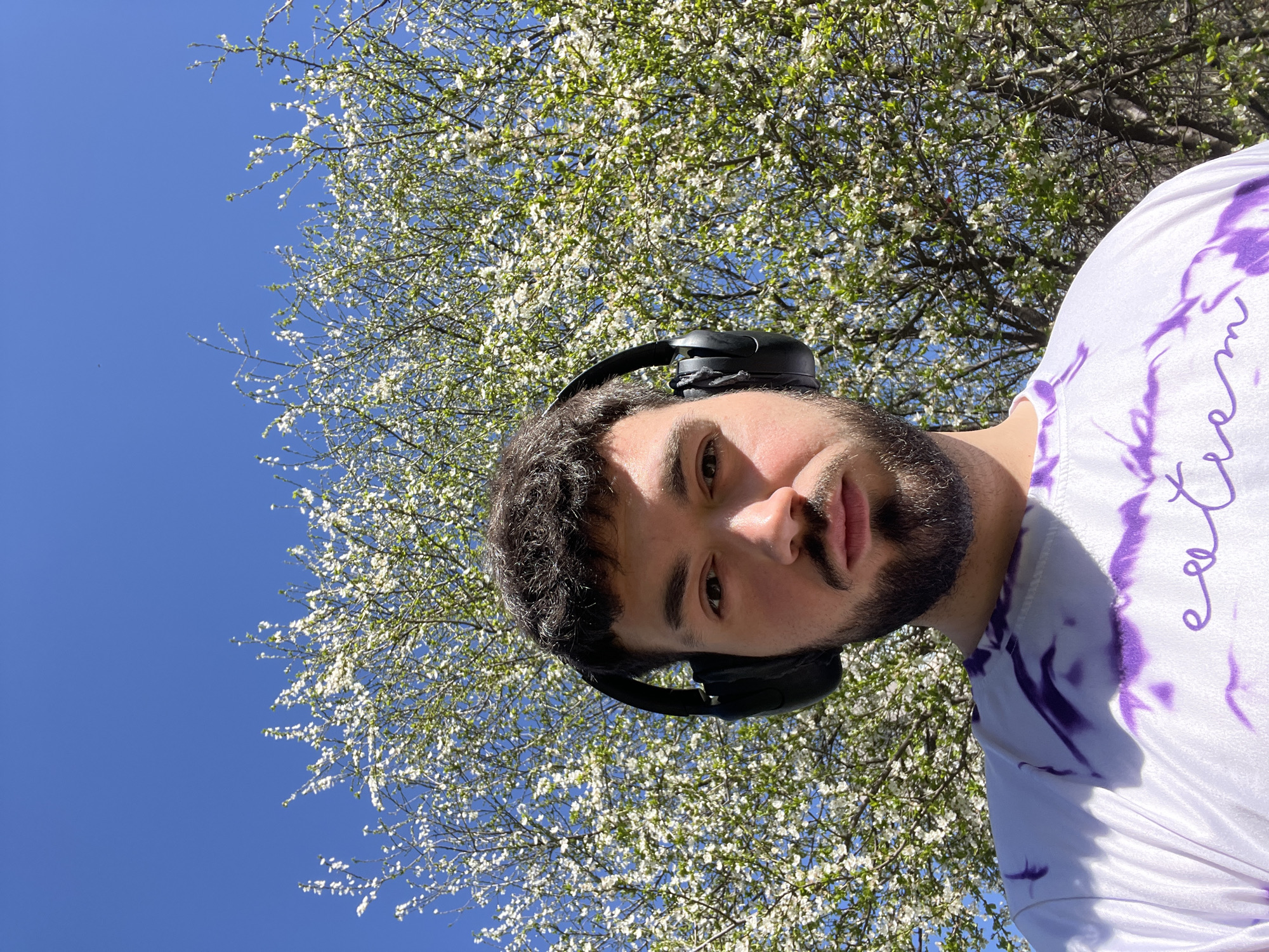 The image shows a selfie taken from a low angle, looking up at a man with dark hair and a beard, wearing black headphones. He is wearing a white t-shirt with purple tie-dye patterns and text. Behind him, there is a tree full of white blossoms against a clear blue sky. Based on the common presence of blooming trees in spring and the architectural style possibly visible in other images, it's plausible that the picture was taken outdoors in a park or green space in Sofia, Bulgaria, although without further architectural details this cannot be stated with certainty.