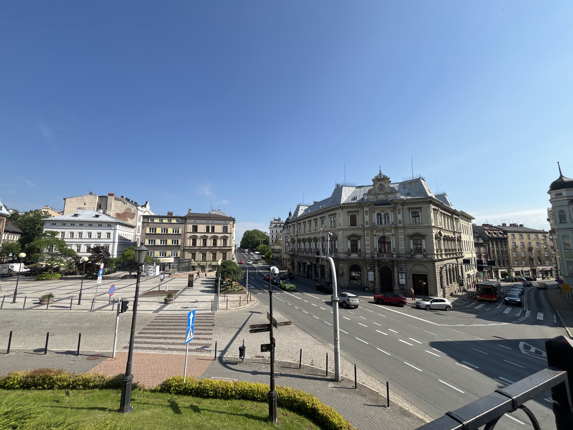 This is a wide-angle, eye-level shot of a city street scene on a clear, sunny day. The sky is a vibrant, cloudless blue. The scene depicts a wide road with multiple lanes of traffic, lined with ornate, multi-story buildings.
On the left side of the image, a large, open plaza area is visible, paved with cobblestones and lined with lampposts. Pedestrian crossings are marked on the plaza. Several buildings face the plaza, with one white building featuring many windows. Greenery and trees are also present in this area.
The main focus of the image is the street and the buildings lining it. A prominent, ornate, light-colored building with intricate architectural details and a mansard roof dominates the right side of the street. Across the street, other buildings of varying architectural styles and colors are visible, some with mansard roofs and others with simpler facades.
Several cars, including a red sedan, a white hatchback, and a bus, are in motion on the road. A few pedestrians are walking on the sidewalks and crossing the street. Traffic lights and street signs are visible throughout the scene.
Notable details include the intricate architecture of the buildings, suggesting a European city. The bright sunlight casts strong shadows, indicating it is likely daytime. The overall mood is one of urban activity and a pleasant, clear day. There is some visible text on street signs, but it is not legible in the image. One sign on the plaza has a blue arrow pointing upwards. A sign on the street pole has directional arrows with text on them.