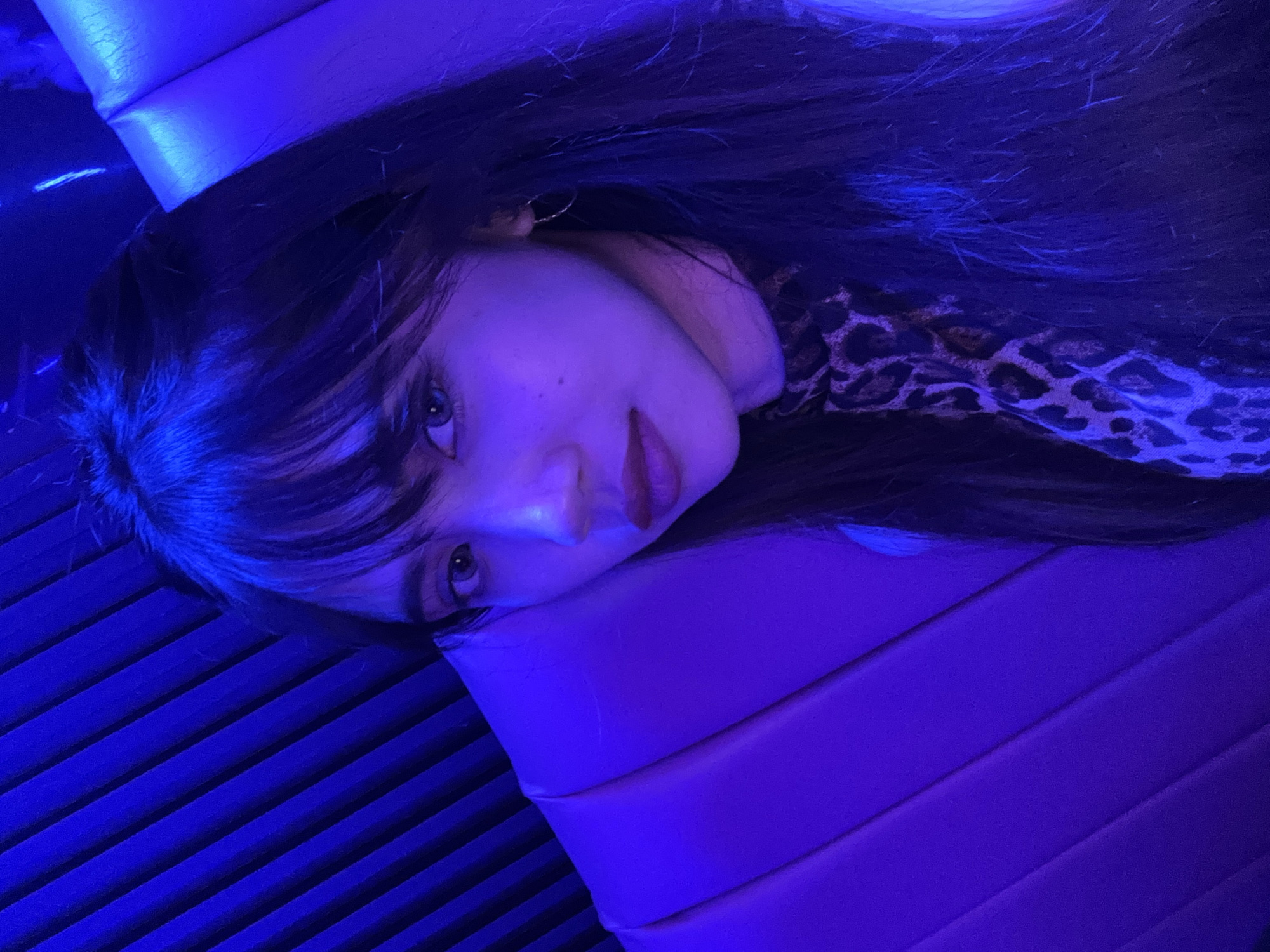 The image shows a close-up, rotated shot of a young woman with dark hair and bangs, lying on her side and looking towards the camera. She is wearing a leopard-print top. Her face is illuminated by a strong blue light, giving the scene a cool and atmospheric feel. The background consists of textured surfaces, appearing to be part of a seating area or a wall with horizontal ridging, also bathed in the blue light.
The woman's expression is ambiguous, with a slight, almost contemplative tilt of her head. Her eyes are open and directed at the viewer, and her lips are slightly parted. The blue lighting casts shadows and highlights, adding a dramatic effect to her features. The overall impression is one of intimacy and perhaps a moment of quiet reflection. There are no visible signs of the time of day or weather. The location context is Davao City, Philippines, but there are no specific visual cues in the image to confirm this. There is no visible text in the image.