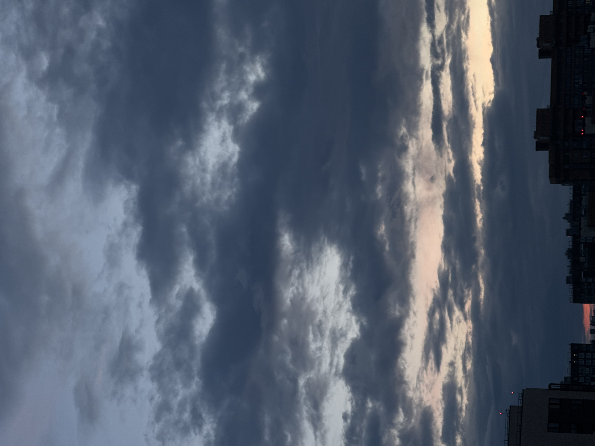 The image depicts a dark, cloudy sky over a cityscape. Buildings are visible at the bottom of the frame, silhouetted against the twilight sky. The clouds are a mix of dark grays and blues, with some lighter patches indicating breaks in the cloud cover or the last vestiges of sunlight. There are no people or distinct actions visible in the image. The environment appears to be urban, likely at dusk or dawn, given the dim lighting and the presence of artificial lights on some buildings. The location is identified as Sinyavino, Russia.