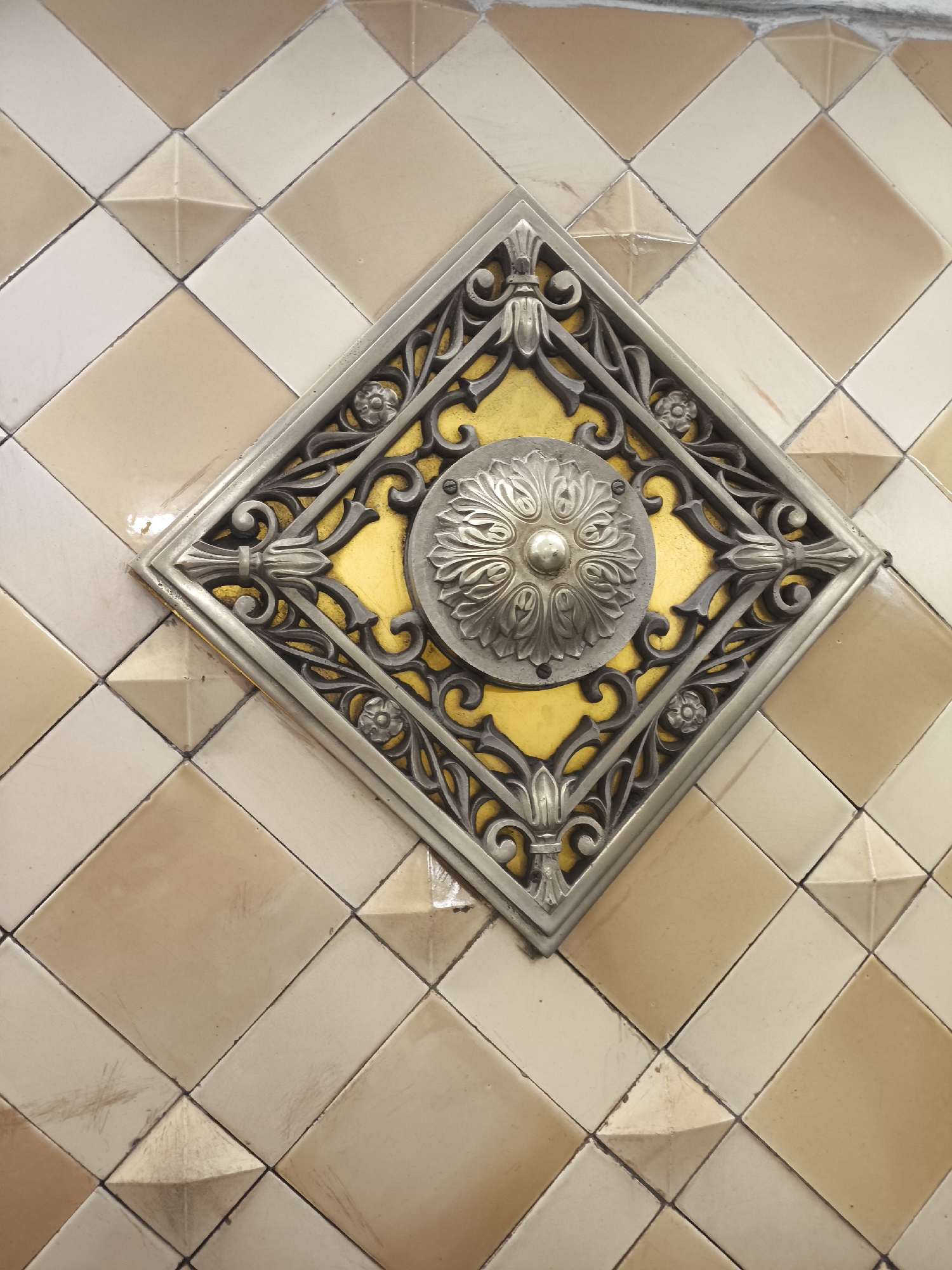A close-up shot of a diamond-shaped decorative piece affixed to a wall composed of diagonally-set square tiles, likely in an indoor setting within the city of Unknown, Unknown. The decorative piece features a yellow-painted background enclosed by a silver metal frame with ornate, floral-like carvings. A silver metal medallion with a radiating floral design is centered on the yellow background. The tiles surrounding the piece are alternately colored in shades of beige and cream, with some tiles having a raised pyramidal center. The tiles are placed in a diamond pattern.