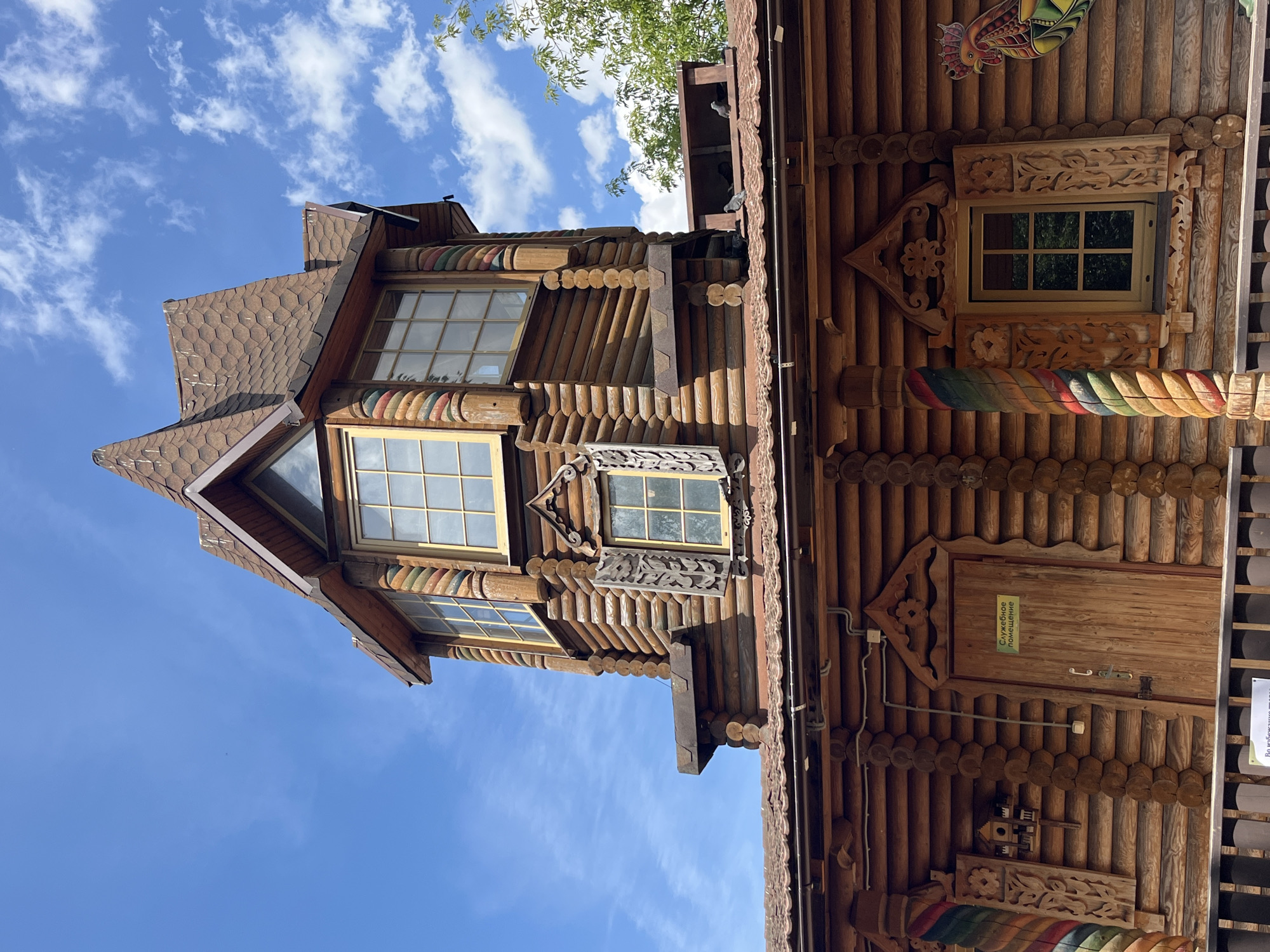 The image shows a portion of a wooden building with a turret-like structure on top. The building is made of logs and features ornate carvings around windows and doorways. The roof is covered with brown shingles. The sky above is blue with scattered white clouds. A green sign with Russian text is visible on a door. There are pigeons on the roof. This structure is part of the "Terem" (Fairy Tale House) at the Children's Museum of Pushkin City Park, located in Pushkin, a municipal town within the federal city of Saint Petersburg, Russia.