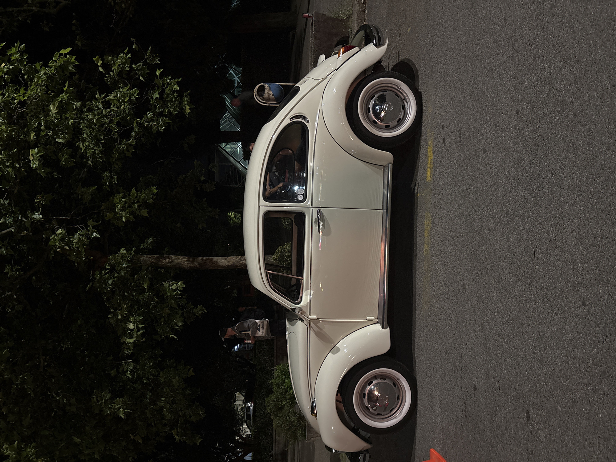 At night in Podgorica, Montenegro, a white vintage Volkswagen Beetle is parked on a street next to a tree. An individual is standing near the car. A building is visible in the background.