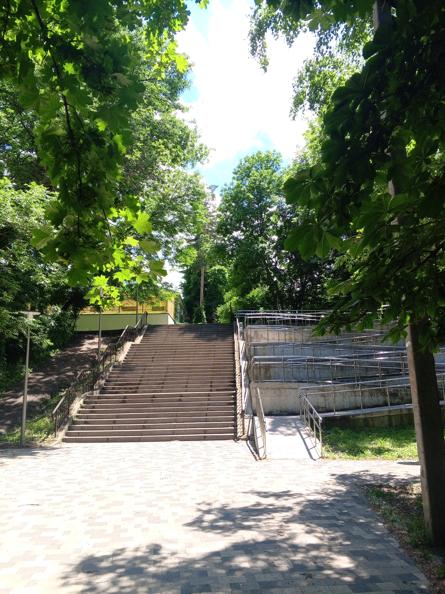 The image depicts an outdoor scene with a large set of concrete stairs leading upwards. To the right of the stairs, there is a tiered concrete structure with metal railings, resembling seating or a viewing area. The area is surrounded by lush green trees and foliage. The ground in front of the stairs is paved with interlocking grey and white tiles, with dappled sunlight casting shadows. A lamppost is visible to the left of the stairs. In the background, a building with a yellow roof and white walls can be seen. The sky above is bright blue with some white clouds. The overall impression is that of a park or recreational area.