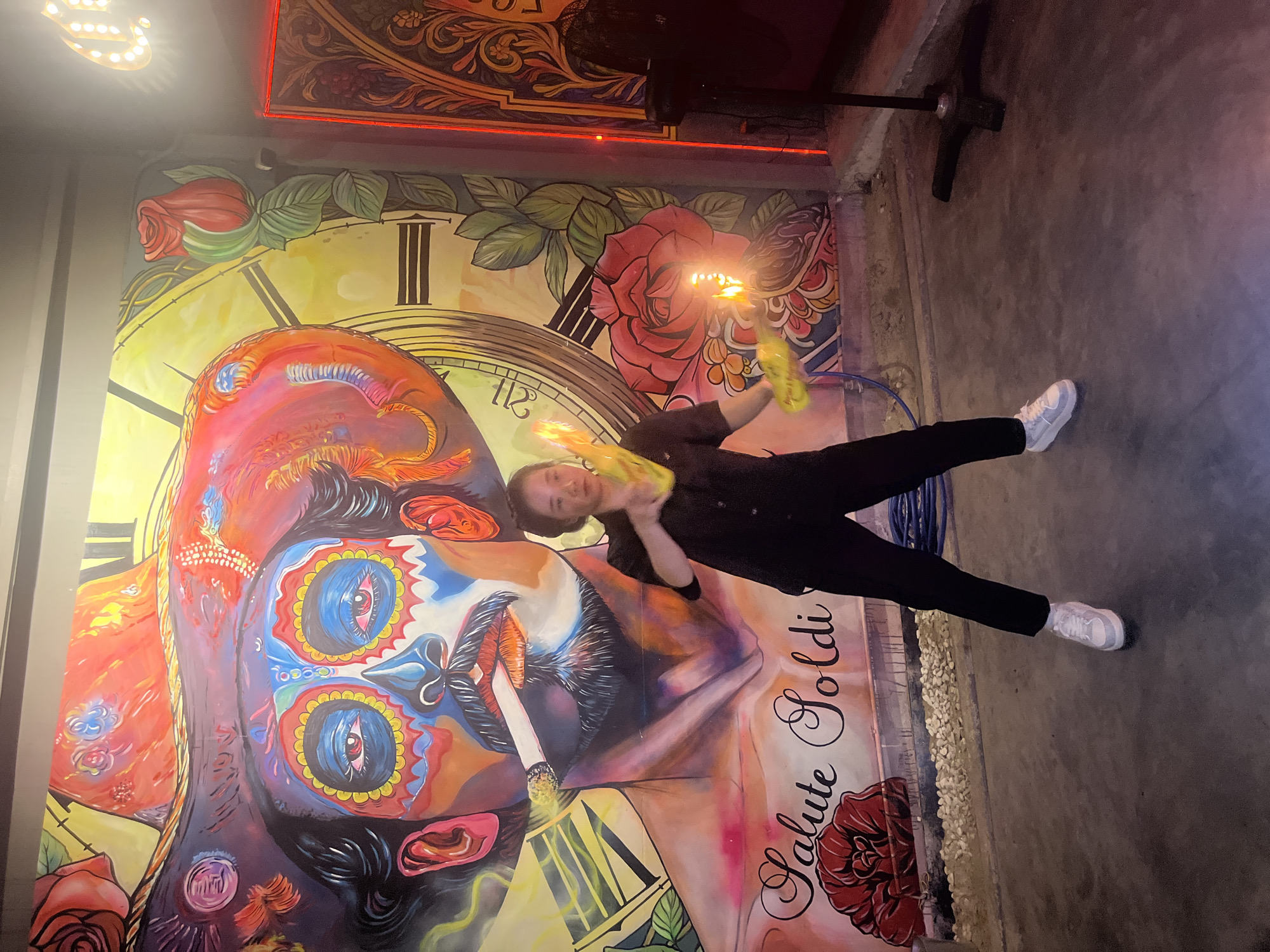 A young person is performing fire tricks in front of a large, colorful mural in Da Nang, Vietnam. The mural depicts a stylized portrait of a man with a sombrero and facial paint, reminiscent of Dia de los Muertos imagery, with a clock face in the background. The person, dressed in a black shirt and pants, is holding two burning props, one in each hand, creating a spectacle of flames. The floor is concrete, and the background shows elements of what appears to be a bar or entertainment venue, including neon lighting and decorative panels.