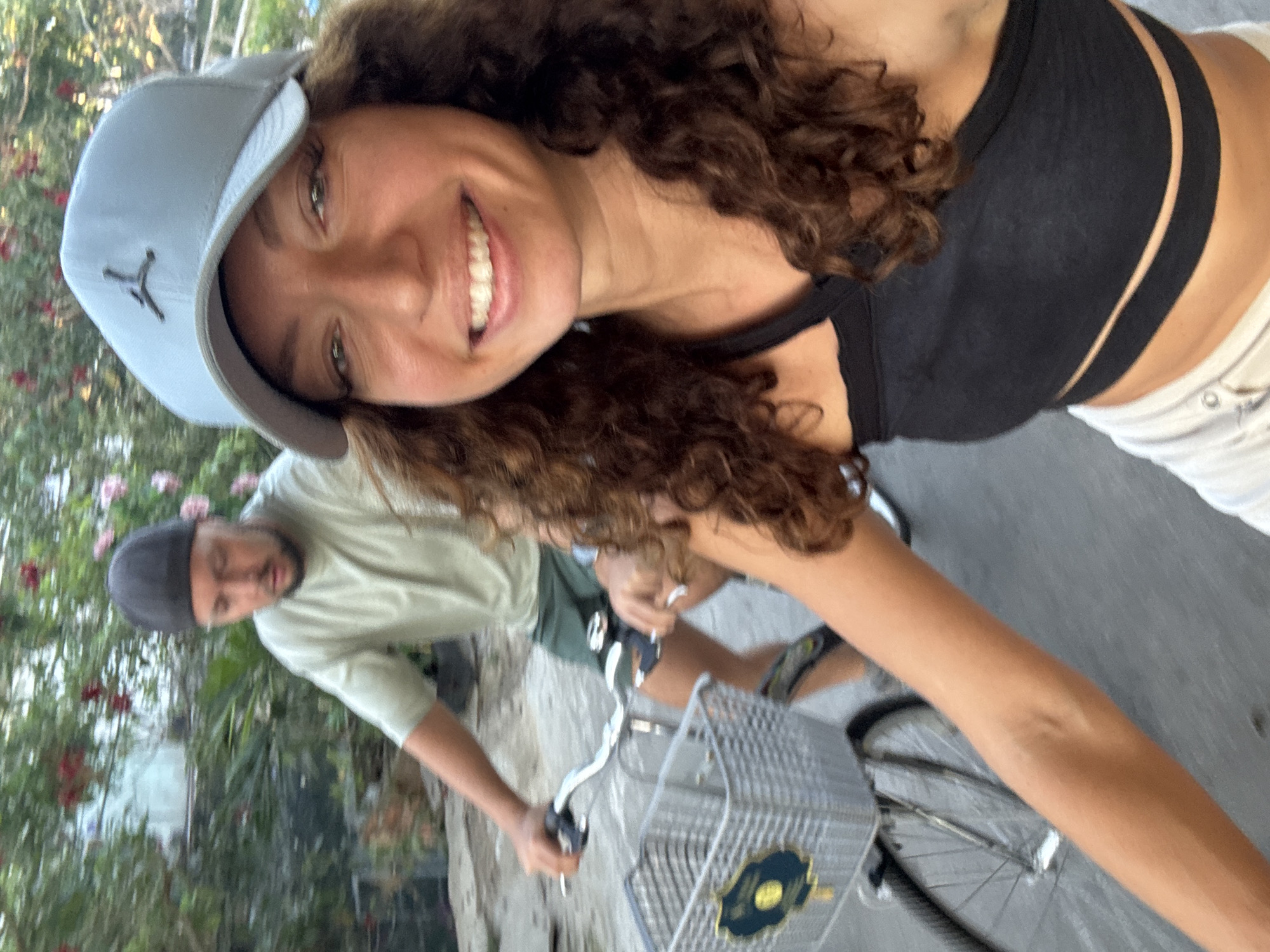 A woman with curly brown hair and a light blue baseball cap smiles at the camera. She wears a black top and white shorts. A man in a green shirt and grey hat stands beside a bicycle. The bicycle has a silver basket. The scene is outdoors with greenery in the background. The location is Hội An Ward, Vietnam.