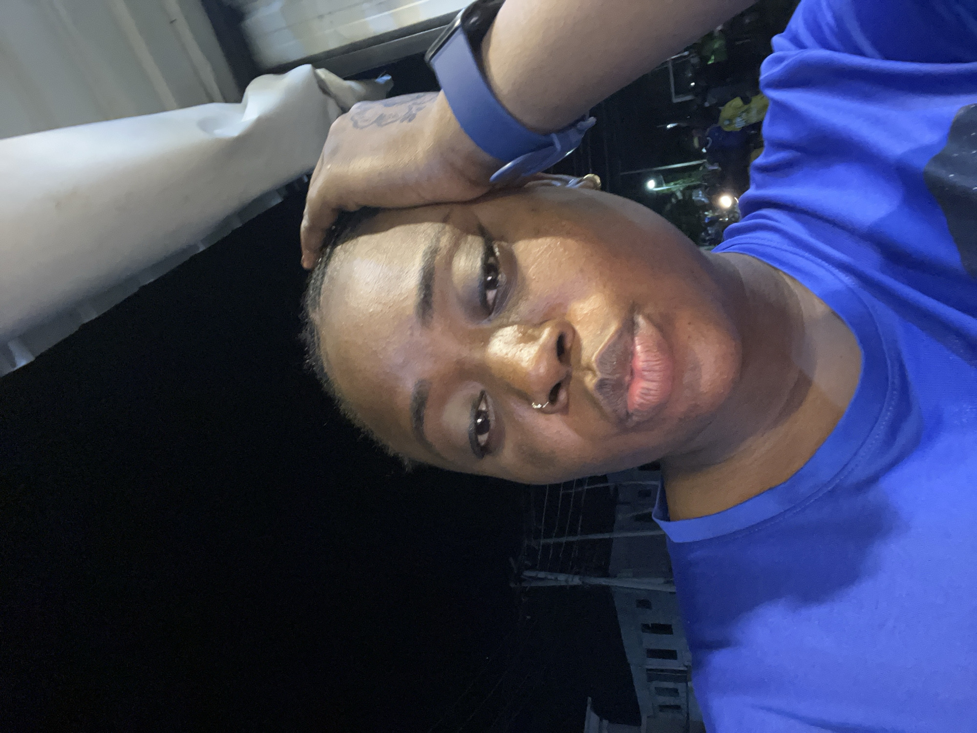 The image shows a person wearing a blue shirt and a blue smartwatch. The person has a nose ring and a tattoo on their hand. They are holding their hand to their forehead. The background is dark, suggesting nighttime, with some blurred lights and structures visible, possibly buildings. The location is Kubwa, Nigeria. The overall scene appears to be an outdoor setting at night.