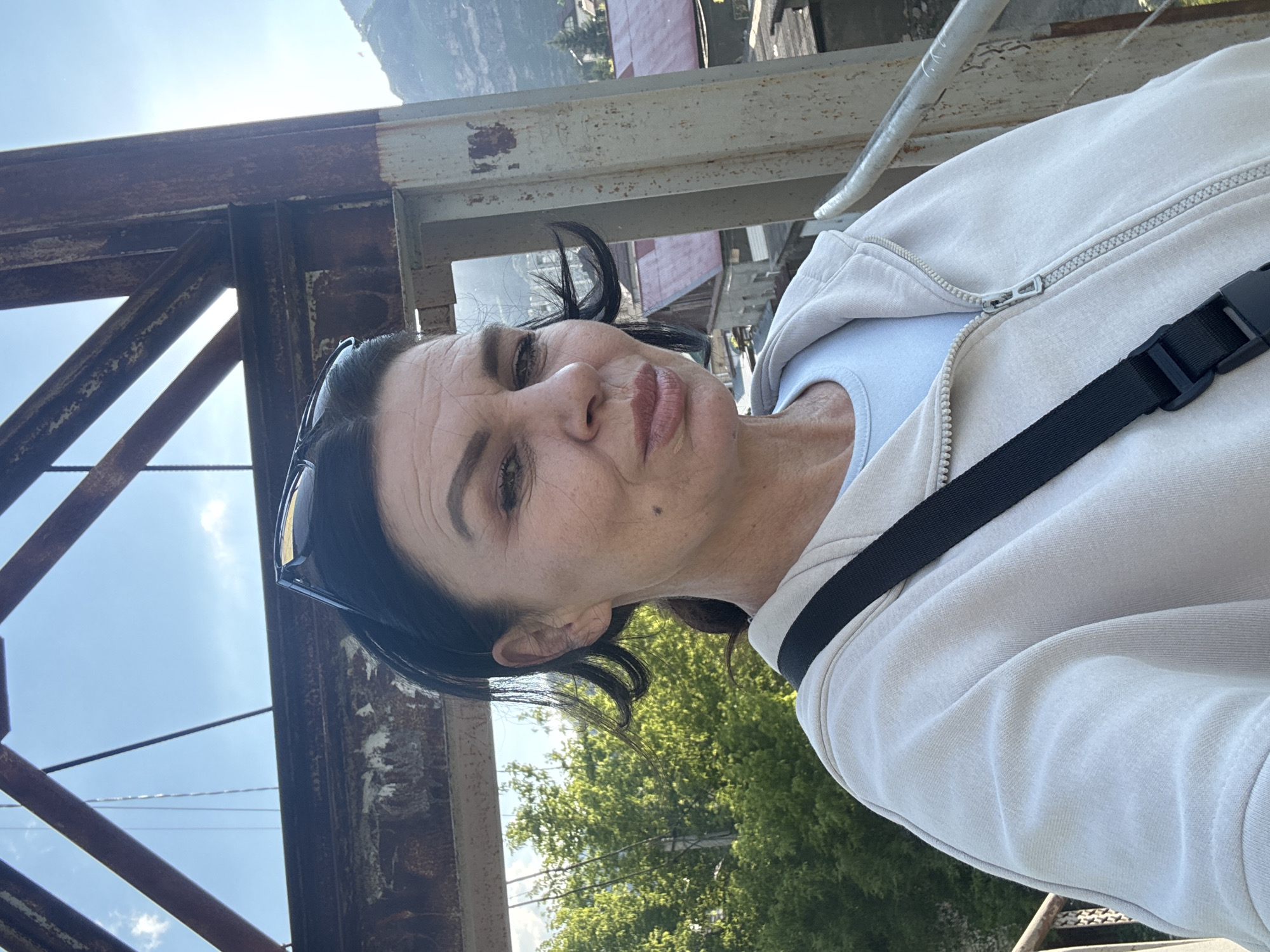A woman with dark hair pulled back and sunglasses on her head is taking a selfie outdoors. She is wearing a light-colored zippered hoodie over a light blue t-shirt and a black strap from a bag is visible across her chest. She is standing on what appears to be a metal structure, possibly a bridge or an overlook, with rusty metal beams and supports visible in the foreground and background. Behind her, there are lush green trees and some buildings with red roofs, suggesting a rural or semi-urban setting within Karachayevsk, Russia. The sky is blue with a few white clouds, and the sun appears to be shining. The woman's expression is neutral, and she is looking towards the camera. The overall impression is a casual outdoor shot captured during the day.