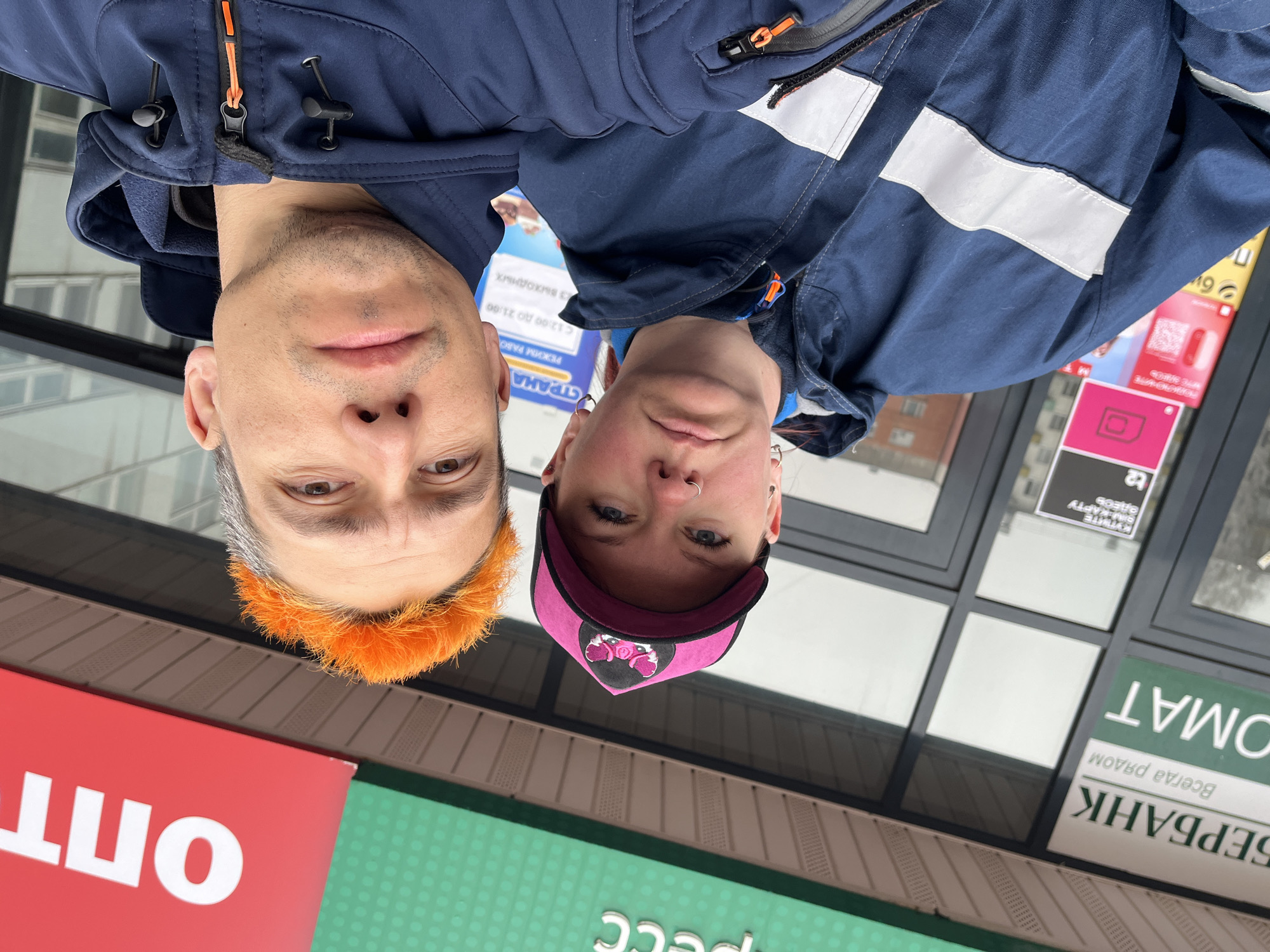 An upside-down, low-angle shot features two individuals, a man and a woman, looking down towards the camera. They appear to be taking a selfie or being photographed from below, outdoors in what seems to be a commercial area of Chaikovsky, Russia.
The man on the left has short, light gray hair with striking bright orange bangs. He wears a dark blue jacket with an orange zipper pull. His expression is neutral.
The woman on the right wears a dark blue jacket with reflective stripes on the shoulders and arm, and a pink baseball cap with a black brim and a light-colored emblem on the front. She has a nose piercing and also displays a neutral expression.
The background reveals a building facade with several signs. Visible Russian text includes "БЕРБАНК" (Sberbank) with its slogan "Всегда рядом" (Always nearby), and a partial "ОМАТ" which is likely part of "БАНКОМАТ" (ATM). Other signs advertise "SIM-КАРТУ" (SIM-card), "СТРАНА" (Country), and "ОПТ" (Wholesale). One sign indicates "РЕЖИМ РАБО" (Operating hours) "С 12:00 ДО 21:00" (From 12:00 to 21:00) with "3 выходных" (3 days off/weekends). The lighting suggests an overcast or cloudy daytime.