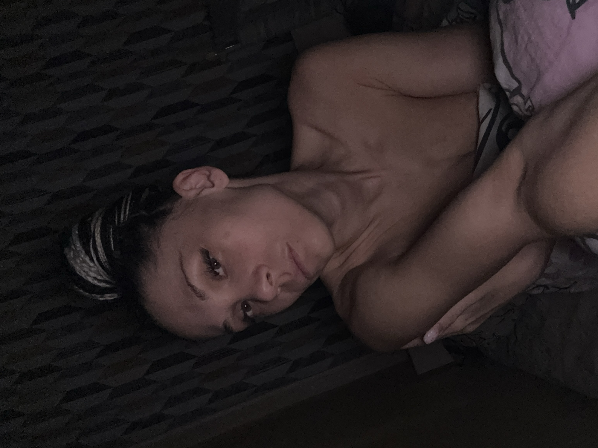 A woman with dark hair with white highlights is lying on a bed in a dimly lit room. She is partially covered by a patterned blanket or sheet. Her face is visible, and she appears to be looking at the camera. The background includes a patterned surface, possibly the bed or a wall. No information is available in the image about the location being Rusanovo, Russia.