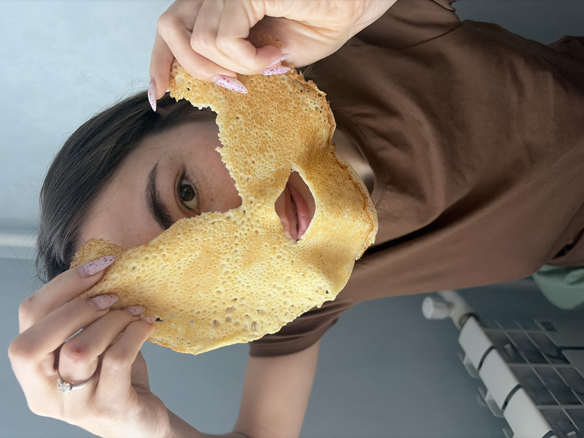 A person is holding a thin, tan, crepe-like food item in front of their face. The food item has holes torn out for the eye and mouth. The person is wearing a brown shirt and has pink and white speckled nails. A ring is visible on their left ring finger. The background is a light blue wall with a white radiator. It is not possible to determine if the location is in Neftekamsk, Russia based on the provided image.