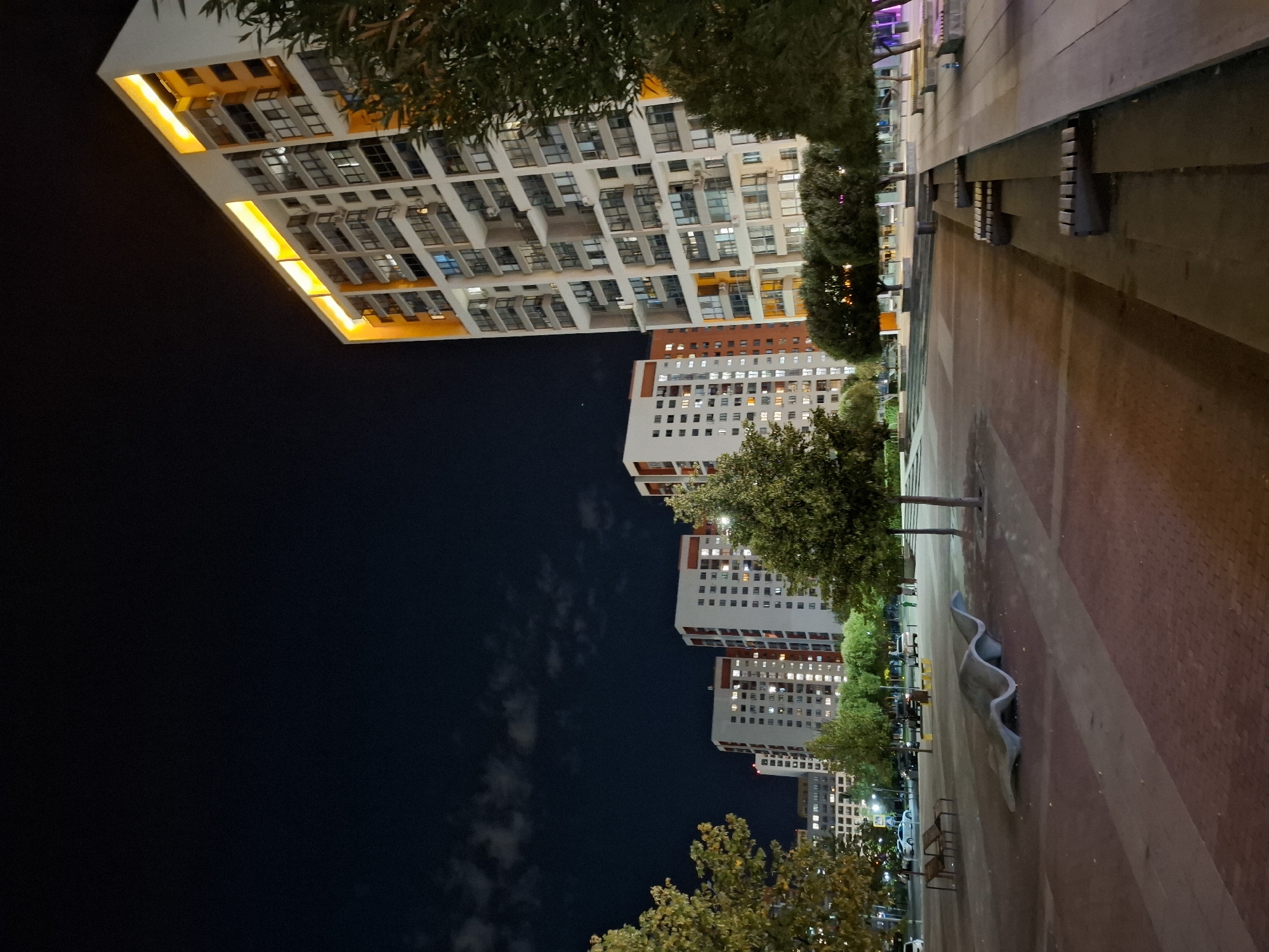 The image depicts a nighttime scene in Tyumen, Russia, featuring a modern residential complex and an outdoor public space. In the foreground, a paved area with a reddish-brown brick pattern is visible, leading to a grey, wavy concrete sculpture. To the right, a wide, elevated wooden platform with benches is illuminated. Behind this platform, several trees are sparsely planted.
Further into the image, a row of trees lines the edge of the paved area. Beyond the trees, a line of multi-story apartment buildings stands, illuminated from within and by external lighting. The buildings are predominantly white with orange accents, featuring numerous windows. The tallest building, on the far right, is brightly lit along its upper edge with yellow lights. In the background, more apartment buildings are visible, some with orange and white facades. A dark night sky with scattered clouds and a few visible lights from the city completes the scene. There are no people or vehicles prominently featured in the image.