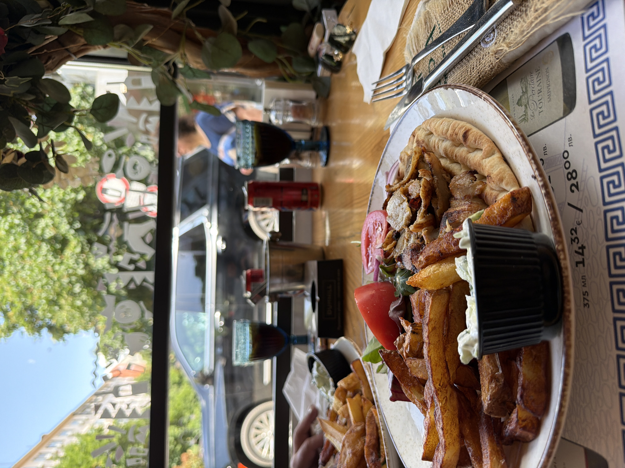 A plate of gyros with fries and tomatoes is centered in the frame on a table in a restaurant, with a container of dip on the plate. The plate sits on a placemat with Greek key designs and text in Cyrillic, next to silverware wrapped in burlap. A bottle of "La Perclaire Touraine Sauvignon" wine is adjacent to the plate. A coca cola can and a blue glass are visible. Through a window, a car and buildings are reflected, suggesting a street scene. Decorative foliage hangs near the window. The location is Sofia, Bulgaria, inferred from Cyrillic text on the placemat.