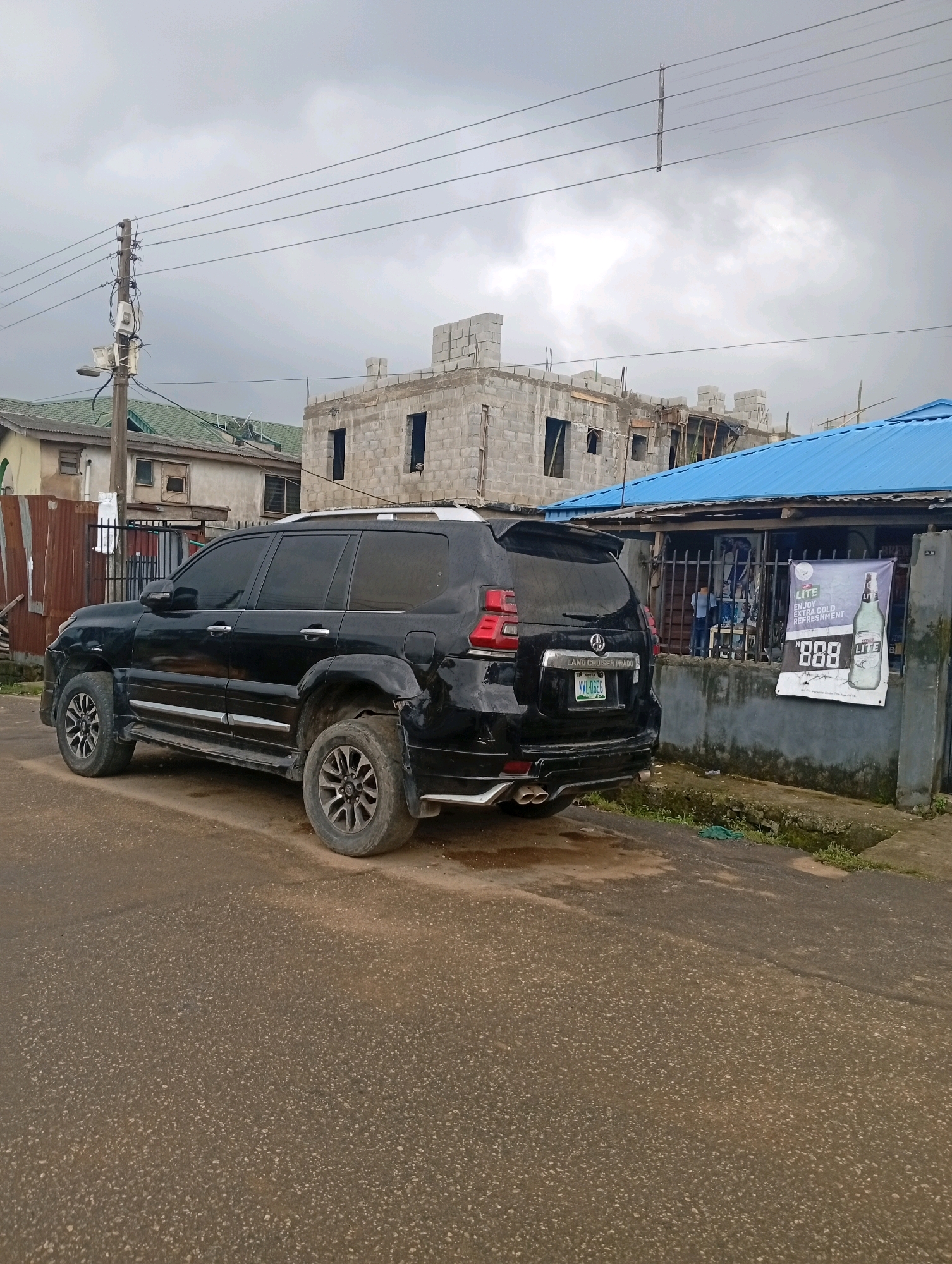 The image depicts a street scene in Akpakpava, Nigeria, under an overcast sky. A black SUV, possibly a Toyota Land Cruiser Prado, is parked on the left side of a paved road. The vehicle exhibits some signs of wear, with visible damage to the rear bumper. In the background, construction is underway on a multi-story building made of concrete blocks. Adjacent to the construction site is a building with a blue corrugated metal roof, appearing to be a commercial establishment, possibly a shop or bar. A banner advertising "Castle Lite" is displayed on the wall of this establishment. Power lines stretch across the upper portion of the frame, supported by a wooden utility pole on the left. Several other buildings with green roofs and metal fencing are visible in the mid-ground. A few individuals are present near the establishment with the blue roof, though their features are indistinct. The environment suggests a developing urban area with ongoing construction and commercial activity.
