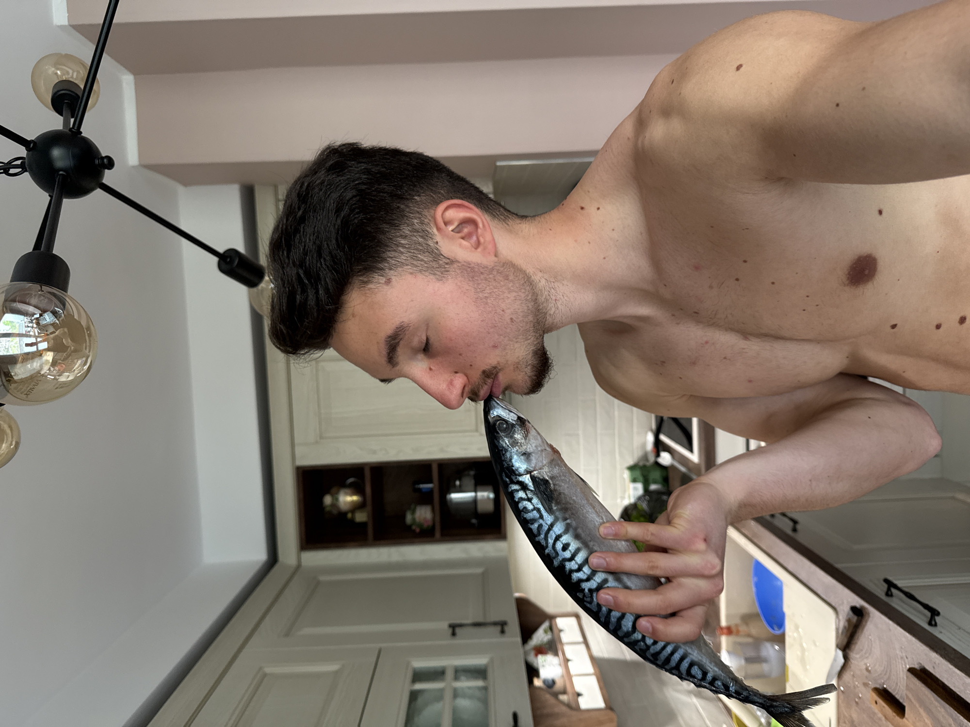 A shirtless man with dark hair and a beard is kissing a mackerel fish. The background shows a kitchen with white cabinets and a modern black chandelier. The location in Sofia, Bulgaria, cannot be determined from the visual information provided.