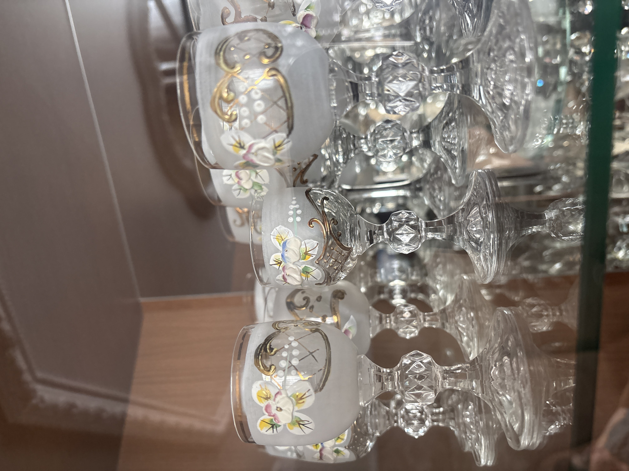 The image displays a close-up, rotated view of decorative glassware, likely part of a display or collection, reflecting on a polished surface. The primary subjects are several small, frosted glass cups or mugs, arranged in rows. Each cup features intricate hand-painted designs: golden, ornate swirls, and delicate white flowers with colorful centers (pink, yellow, light blue, green) accented by small white dots. The cups have clear glass handles.
Behind and around these frosted cups, numerous clear, faceted glass stems and bases of what appear to be wine glasses or other stemmed drinkware are visible. These clear glass elements reflect and refract light, creating sparkling patterns and depth within the composition. The entire arrangement is mirrored on a glossy surface below, showing inverted reflections of the glassware.
A distinct vertical green line is present on the far right edge, suggesting the presence of a glass shelf or divider within a display cabinet. The background reflects muted brown and beige tones, possibly from a wall or furniture in the room. There is no discernible activity taking place, nor are there any people present. No text is visible. The scene is brightly lit, indicative of an indoor setting, with no specific time of day or weather conditions evident. The overall impression is one of delicate and elegant glassware artfully arranged.