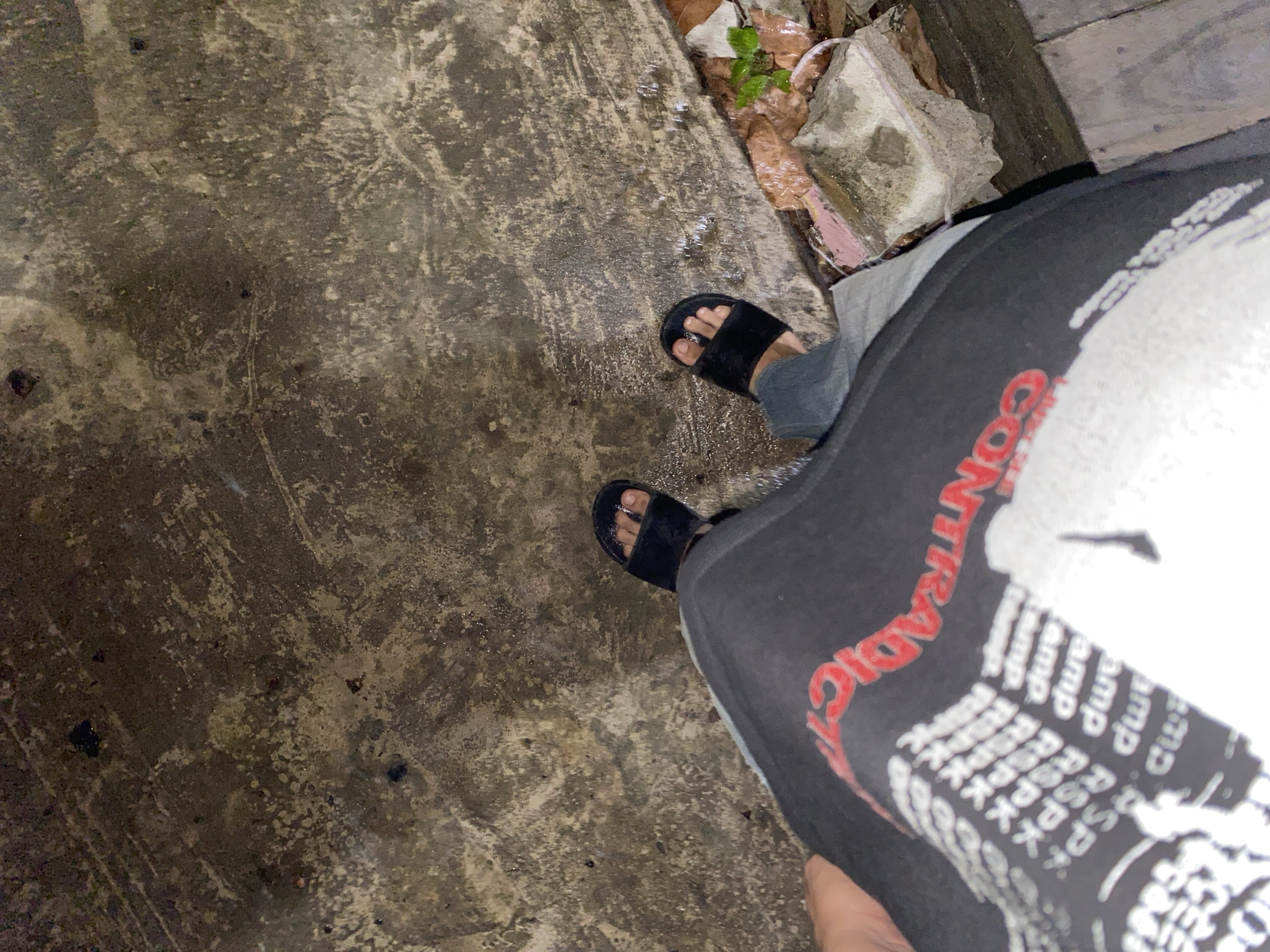 The image shows a person's feet wearing black slides, standing on a wet, textured concrete surface. The person is wearing dark pants and a black t-shirt with red and white text. Some debris, including leaves and a piece of pink insulation, is visible on the ground to the right. The scene appears to be outdoors, possibly a street or walkway in Banjarmasin, Indonesia, indicated by the presence of wet concrete and natural elements.