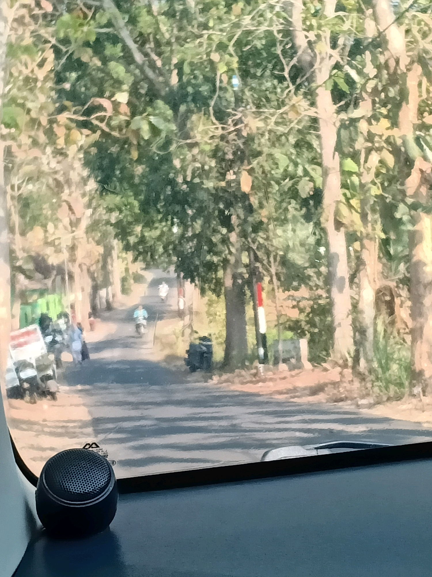 The image depicts a street scene viewed from inside a vehicle, likely in Sucolor, Indonesia. The road is lined with tall trees, creating a canopy over the asphalt. Several people are visible on the left side of the road, standing near what appear to be motorcycles and a cart. Two individuals are on motorcycles, one in the middle distance and another further down the road, both moving away from the viewer. A vertical pole with red and white stripes stands near the right side of the road. The foreground shows the interior of the vehicle, with a dark blue dashboard and a small, circular black speaker. The overall impression is of a sunny day on a rural or suburban road.