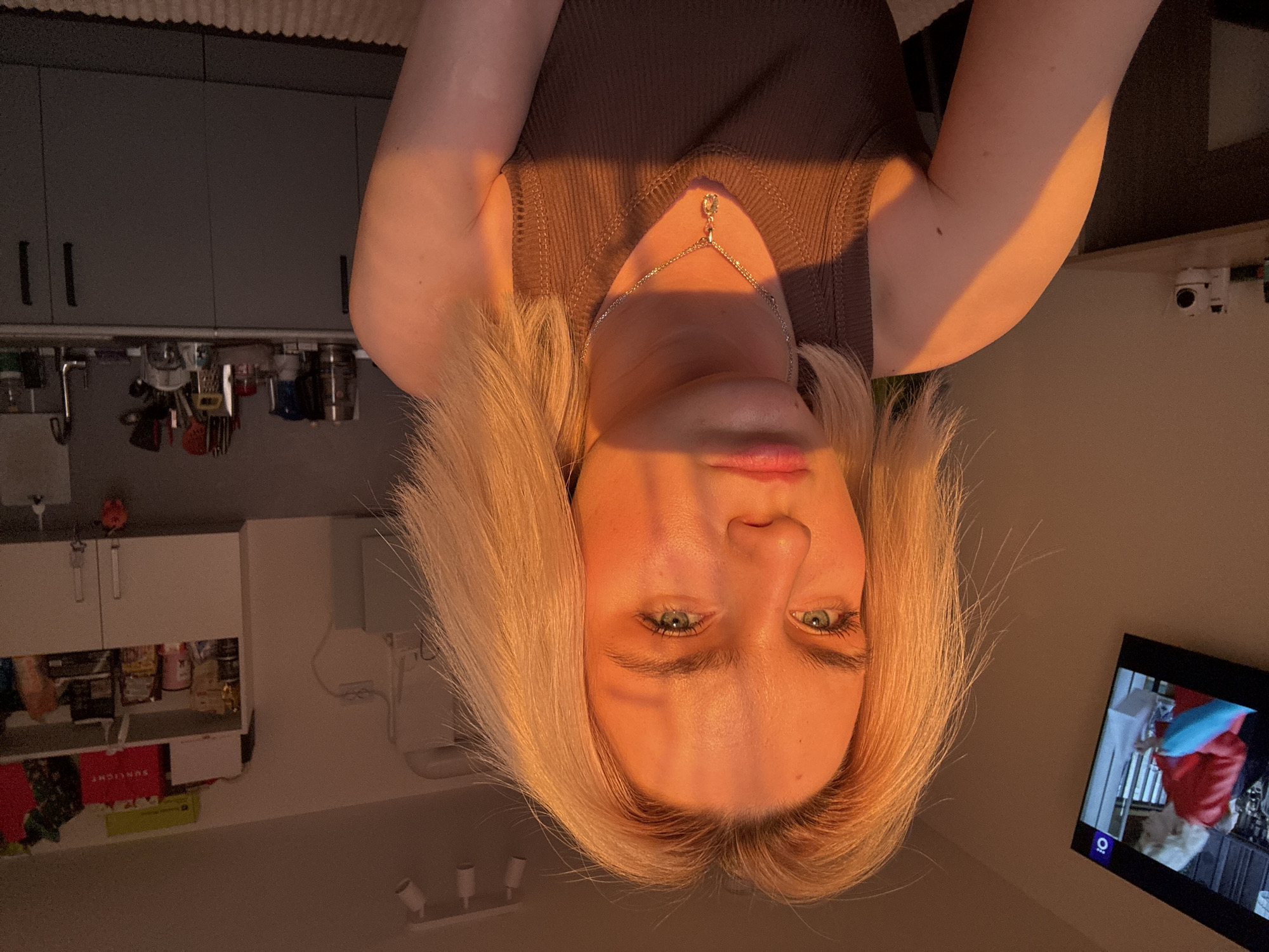A blonde-haired woman with fair skin and green eyes is looking directly at the camera. She is wearing a brown tank top and a silver necklace. The room appears to be a kitchen, with cabinets and appliances visible in the background. A television is on in the corner, displaying an image. The photograph is taken indoors under warm lighting conditions.
I cannot provide the location in the city of Vsevolozhsk, Russia. The image does not contain the necessary information for it to be placed in that specific location.