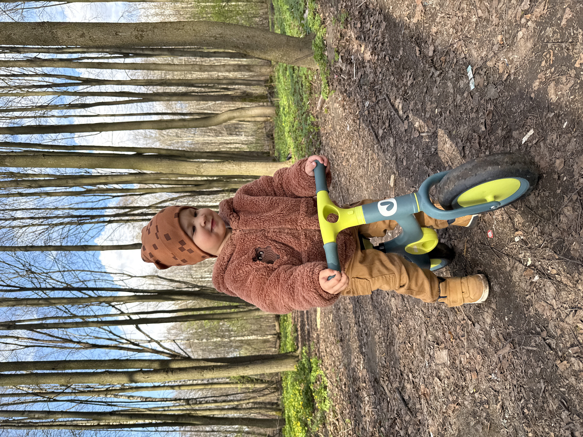 A young child stands with a balance bike on a dirt path in a wooded area, possibly Rusanovo, Russia. The child is wearing a brown knit cap, a brown teddy-bear jacket, and tan pants and boots. The balance bike is predominantly blue-green with yellow accents. The path is covered with leaves and debris, and trees are visible in the background, with glimpses of the sky between their branches.