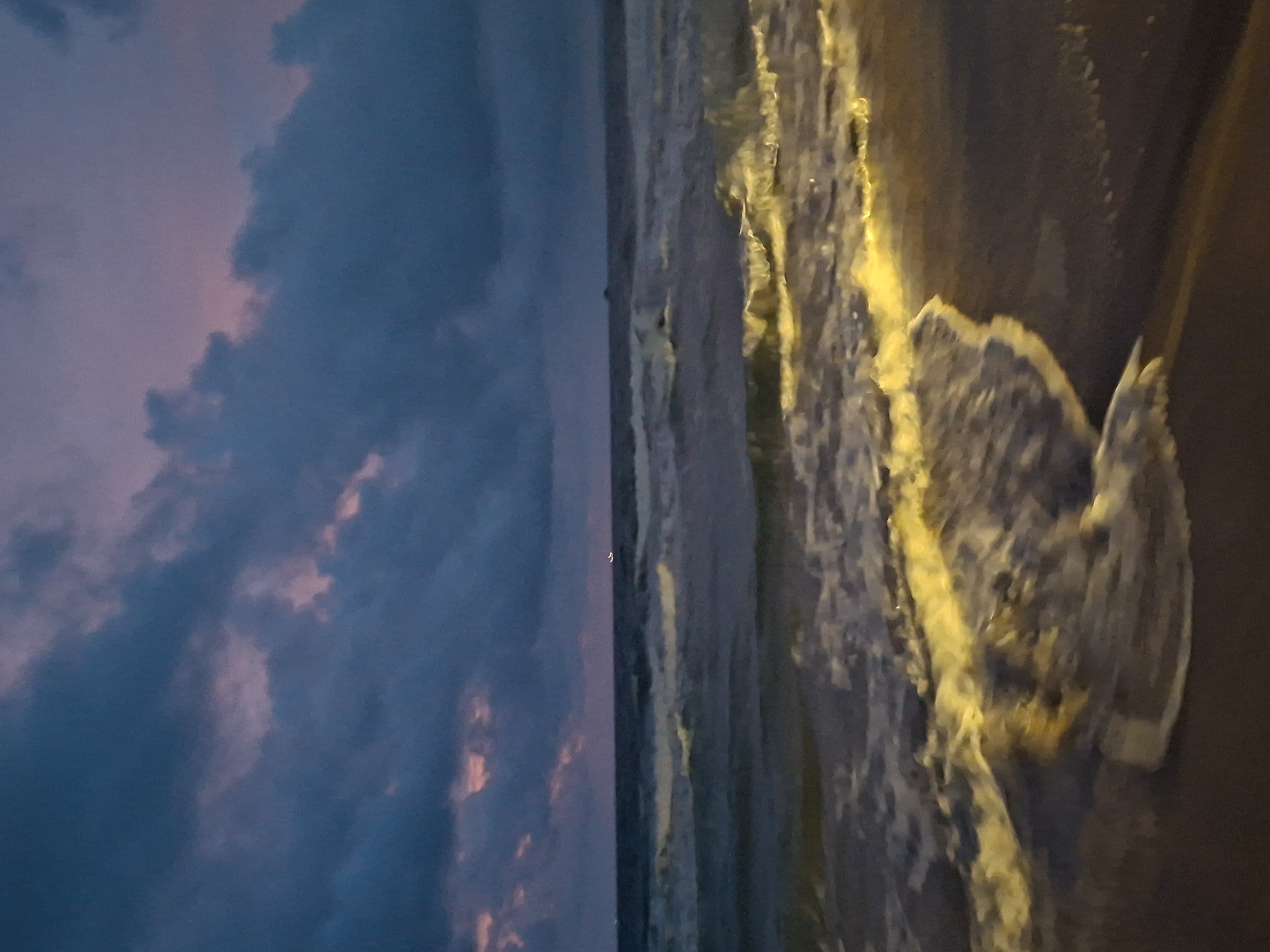 This is a photograph of a beach at dusk, likely taken in Phú Quốc, Vietnam. The sky is filled with dark, dramatic clouds tinged with purple and pink hues, suggesting the sun has recently set or is about to rise. The ocean is a deep blue-grey, with waves gently rolling towards the shore. The crests of the waves are illuminated by a warm, golden light, creating a striking contrast with the dark water and sky. The sand in the foreground is dark and wet from the receding tide. In the distance, a faint light is visible on the horizon, possibly from a boat or a structure. The overall impression is one of tranquility and natural beauty at the transition between day and night.
