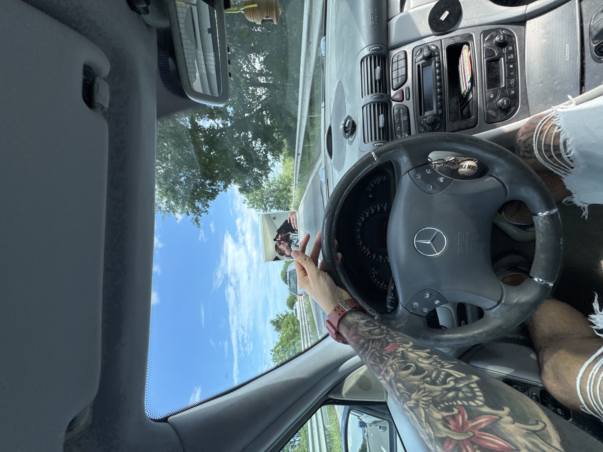 The image presents a first-person view from the driver's seat of a Mercedes-Benz vehicle, positioned on a highway in the city of Burgas, Bulgaria, during daytime under a clear blue sky with scattered white clouds. The driver's left forearm, extensively covered with dark, intricate tattoos depicting various patterns including red floral elements, rests on the steering wheel. A reddish-brown digital watch is worn on the left wrist. The driver's right hand lightly grips the top portion of the grey Mercedes-Benz steering wheel, which features the brand's emblem and integrated controls. A keyring with multiple keys and a "SAN FR" keychain hangs from the steering column.
The grey dashboard displays an instrument cluster indicating a speed between 80 and 90 (likely km/h). The central console contains an infotainment system, air vents, climate controls, and an emergency flasher button. A small black circular object is affixed to the dashboard near the windshield. The driver's knee, covered by light-wash denim jeans with horizontal rips, is visible in the lower left quadrant.
Through the front windshield, a multi-lane highway is visible, bordered by green trees and a metal guardrail. A white truck directly ahead displays a large advertisement on its rear, featuring two individuals, a man and a woman, in a close embrace. The car's interior rearview mirror is visible in the upper right, with a small wooden, bell-shaped air freshener hanging from it. The grey interior A-pillar and roof lining are also partially visible. The driver's side exterior mirror partially reflects the road and trailing vehicles.