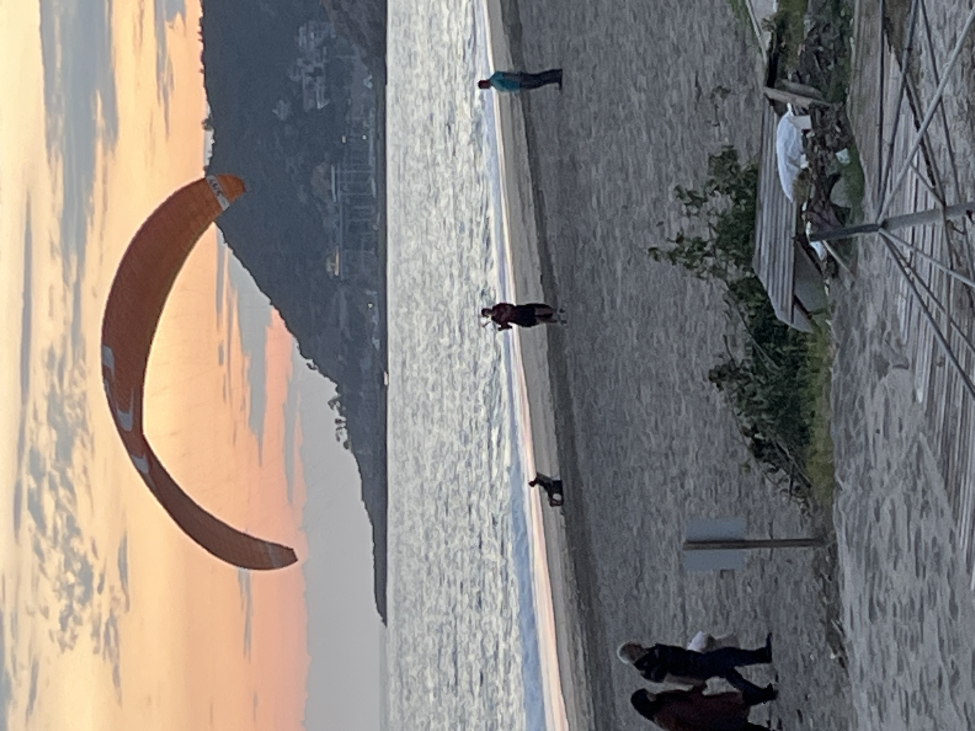 The image depicts a beach scene at Alanya, Turkey. The horizon is vertically oriented with the sky on the left and the beach on the right. In the sky, an orange kite surfing parachute is inflated and partially visible. The sky behind it exhibits a mix of light orange, grey, and white hues, indicative of sunset.
The beach is sandy, and the sea appears calm. Several individuals are present on the beach at various distances from the camera. An individual in a light blue shirt stands in the water. Nearer, a person in dark clothes is visible at the water's edge. A dog is running along the beach between these two people.
In the foreground, two people are standing on the sand. One is wearing a dark jacket and has grey hair. The other is in a maroon jacket. A signpost is next to them.
Part of a boardwalk with an umbrella is situated to the right of the frame.
The background on the left shows buildings on a hillside.