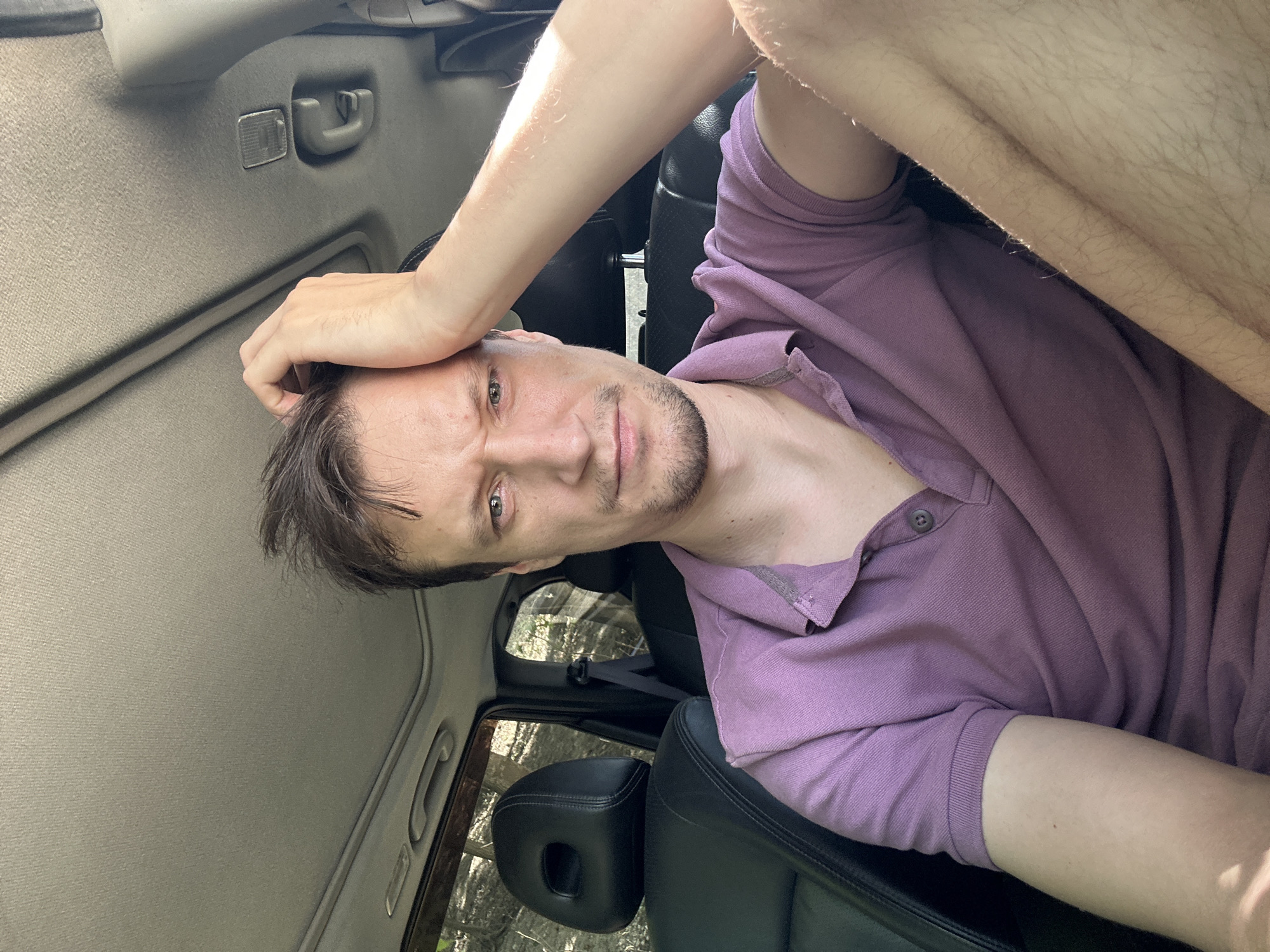 In the image, a man is seated inside a car, looking directly at the camera. He has light skin, brown hair, and is wearing a purple polo shirt. His right hand is raised to his head, touching his hair. The car interior is visible, including black leather seats and the sunroof. Outside the car windows, trees and foliage suggest a natural environment, possibly a rural or wooded area, which could be consistent with the outskirts of Makiv, Ukraine. The lighting indicates it is daytime.