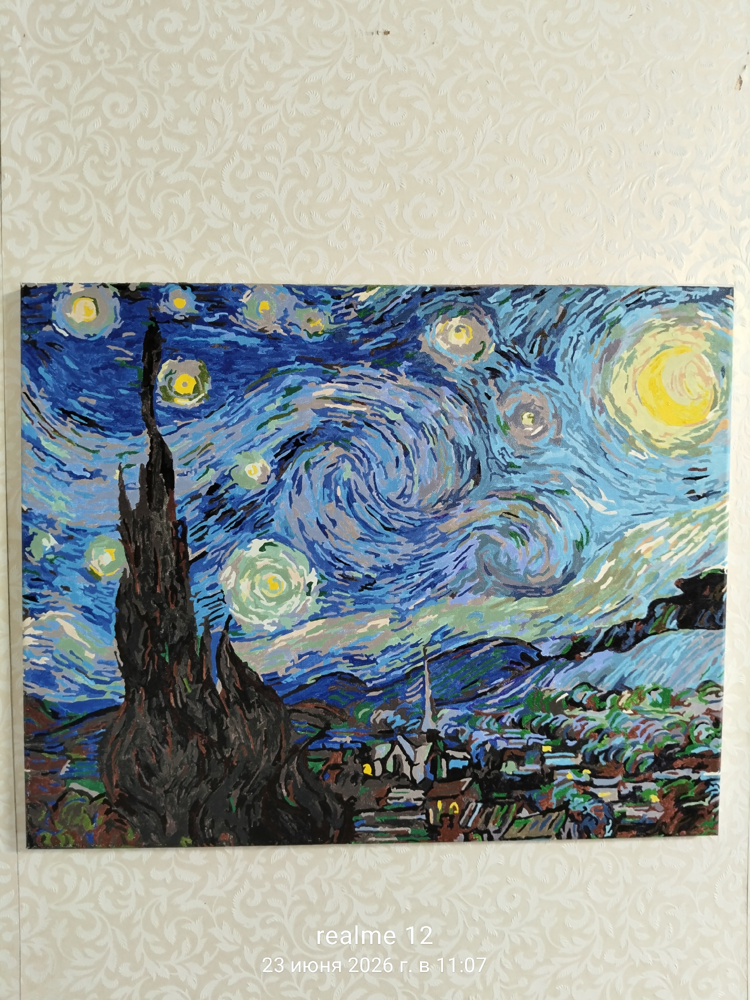 The image displays a painting, likely a recreation of Vincent van Gogh's "The Starry Night," mounted on a wall with textured wallpaper. The painting depicts a swirling night sky with prominent stars and a crescent moon, a dark, flame-like cypress tree in the foreground, and a village with illuminated windows below rolling hills. The painting is positioned centrally on the wall, with the wallpaper creating a patterned background. The bottom of the image shows a timestamp overlay reading "realme 12 23 июня 2026 г. в 11:07", indicating the image was captured on June 23, 2026, at 11:07 AM. There are no people or discernible actions taking place within the scene of the painting itself. The location of Shchyolkovo, Russia is not visually represented in the image; it is information external to the visual content.